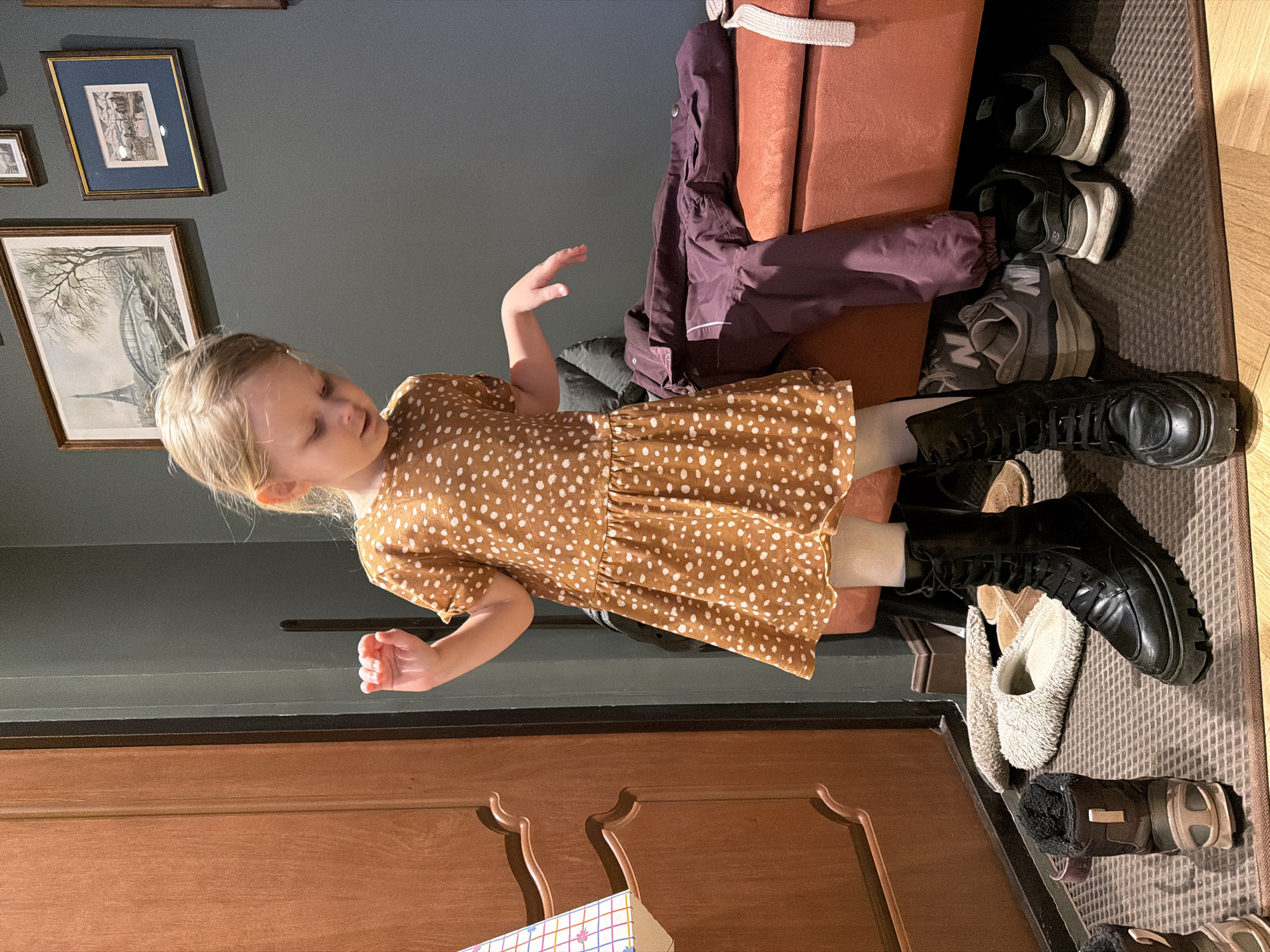A young girl with blonde hair stands in an entryway. She wears a brown dress with white polka dots and white tights, and large black boots. Her arms are raised. Behind her is a padded bench with a brown jacket draped over it. Various pairs of shoes are arranged on the floor. There are framed pictures hanging on a dark grey wall behind the girl. A brown wooden door is to the left of the girl. The location is likely a home interior. There is no information in the image to confirm it is in Khimki, Russia.