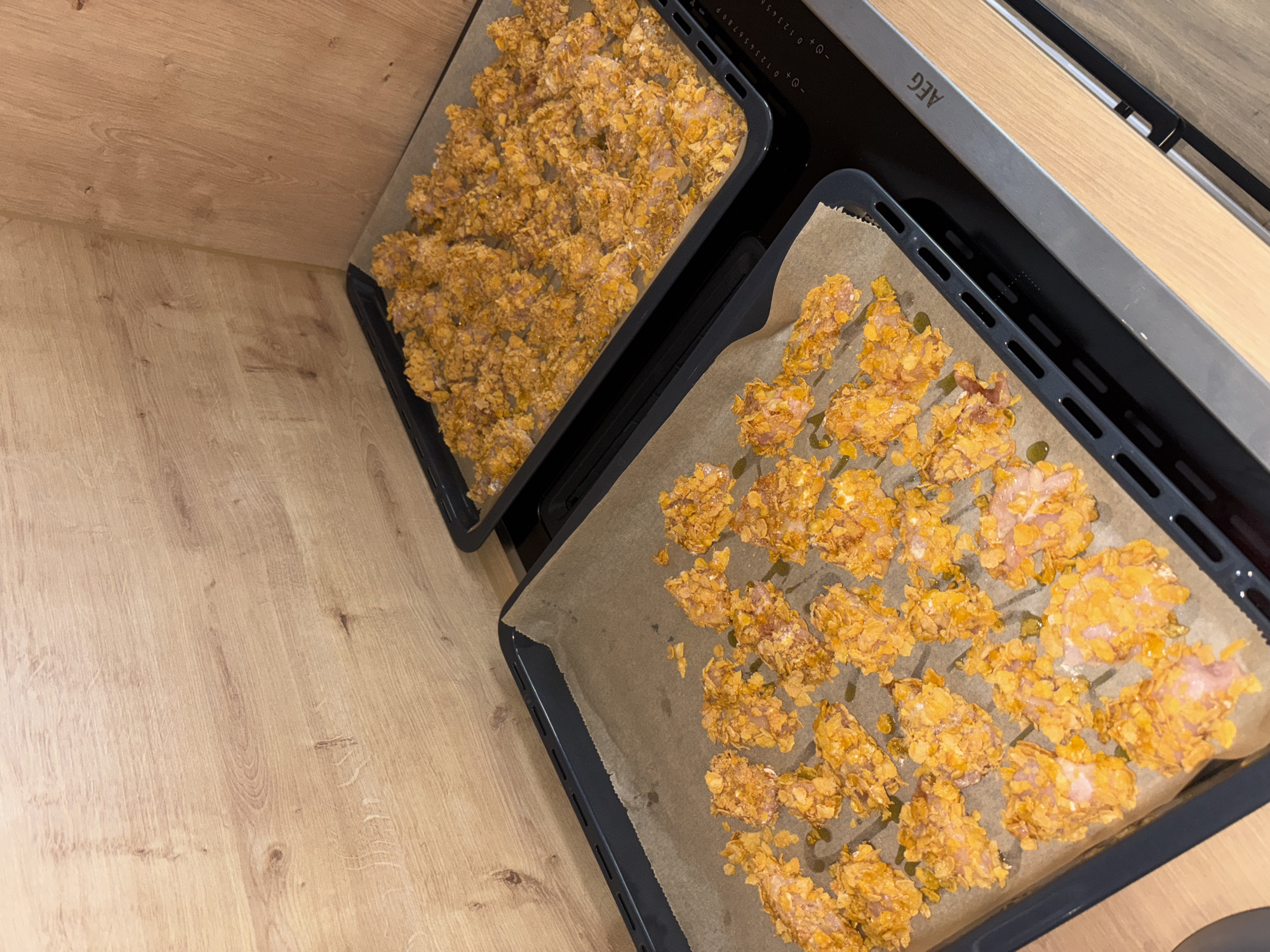 The image shows two baking sheets filled with food items, likely chicken tenders, coated in crushed cereal, possibly cornflakes, and seasoned with spices. The baking sheets are placed on a stovetop in a kitchen. The background features a wooden-paneled wall. The stovetop is an AEG brand, with visible control knobs and markings. One baking sheet is closer to the foreground and contains individual pieces of the coated food, arranged in rows. The second baking sheet is further back and contains a larger, more densely packed portion of the same food. Some of the food pieces appear to have a light drizzle of oil on them. The lighting suggests an indoor setting, possibly during the day or under artificial kitchen lights. No people are visible in the image. The scene depicts food preparation, specifically the arrangement of coated food items on baking sheets, ready for cooking. The location is not specified as Viernheim, Germany in the image content.