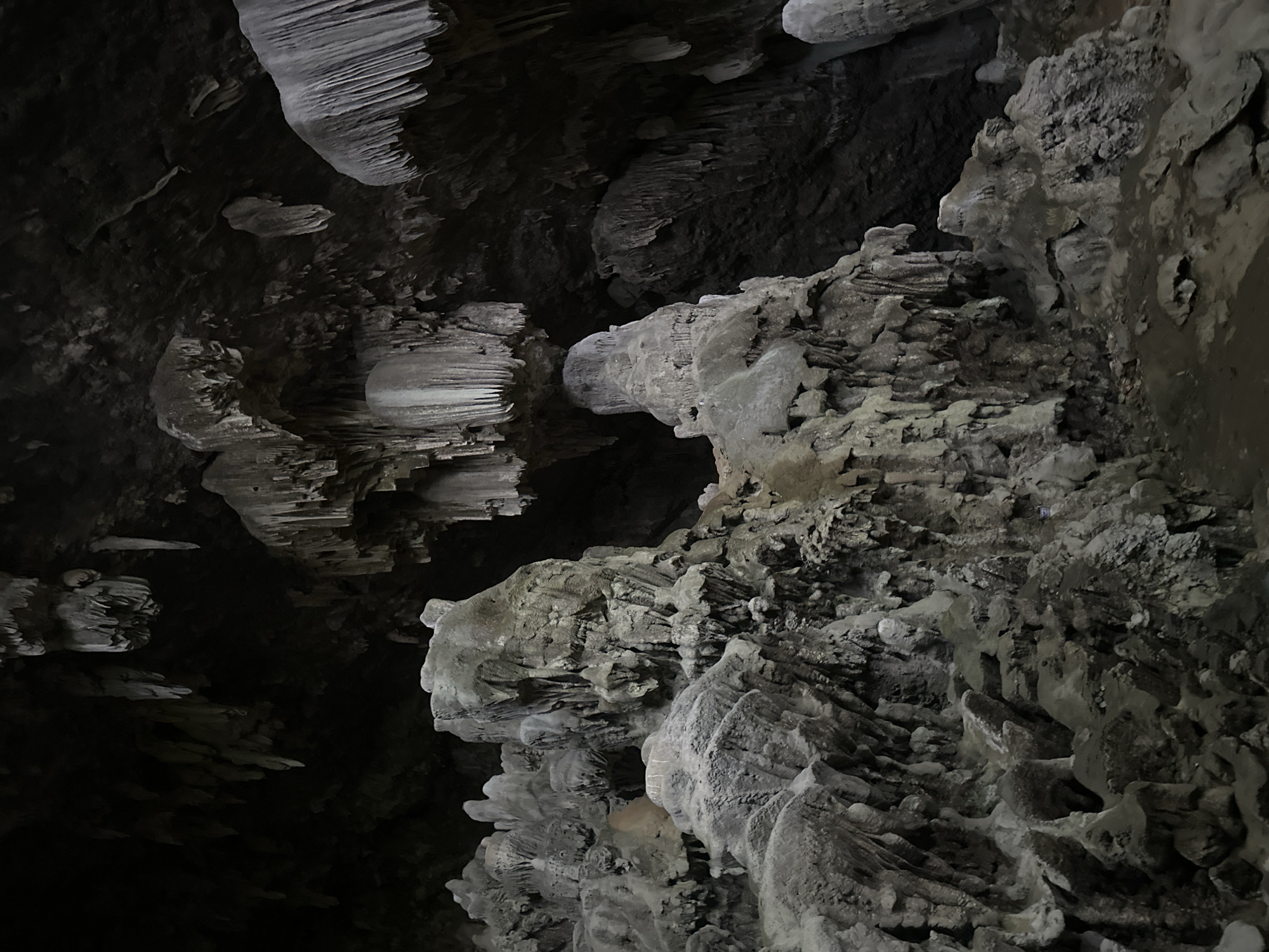This image depicts the interior of a cave in Nà Mèo, Vietnam. The scene is dominated by natural geological formations, specifically stalactites hanging from the cave ceiling and stalagmites rising from the cave floor. These formations are varied in shape and size, with some appearing smooth and column-like, while others are more jagged and textured. The cave's ceiling is dark and appears rough-hewn, with numerous stalactites of varying lengths suspended from it. The floor of the cave is also visible, covered in rocky debris and with a path winding through the formations. The lighting is dim, casting long shadows and highlighting the textures of the rock. No people or animals are visible in the image. The overall environment is one of natural erosion and mineral deposition, creating a complex and ancient subterranean landscape.