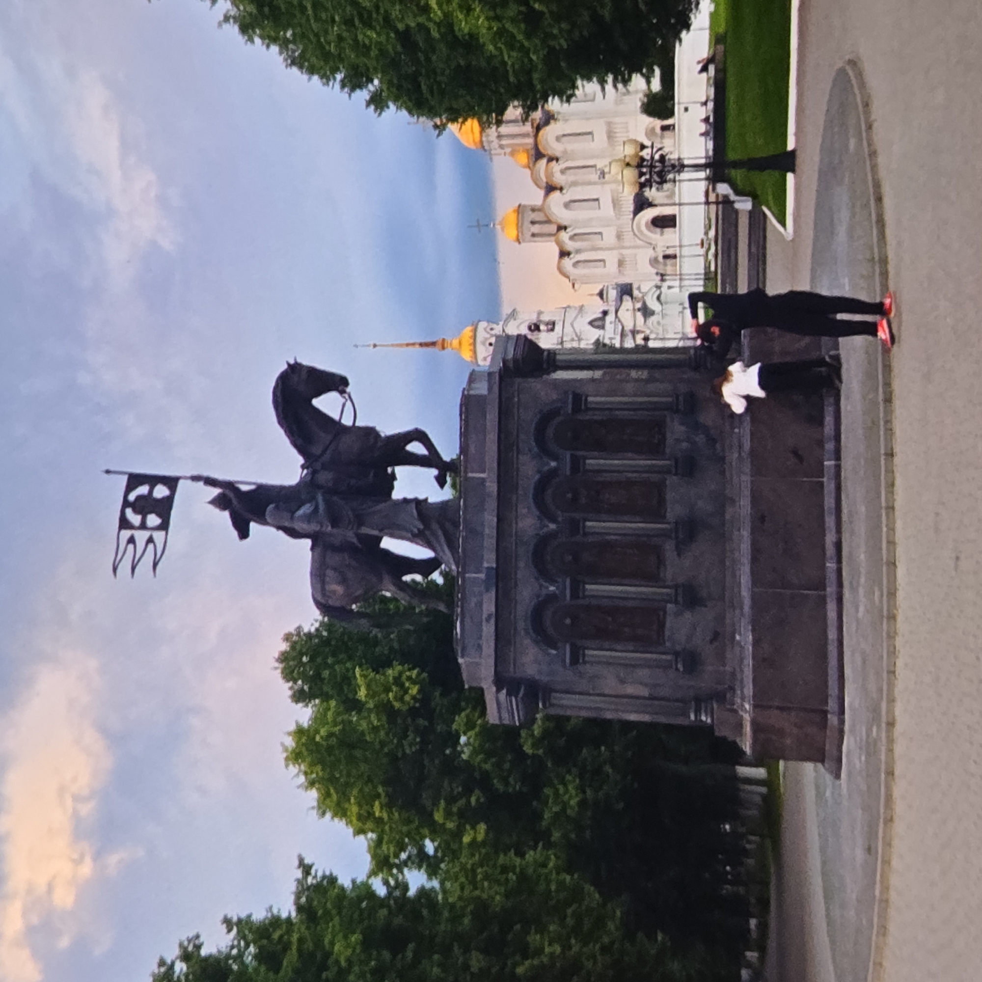 An equestrian statue stands prominently in the foreground of the image, with the figures of two people visible near its base. The statue depicts a figure on horseback holding a flag with a distinctive emblem. Behind the statue, a white church with golden domes and a tall spire is visible, its architecture suggesting a religious building. Lush green trees frame the scene on both sides, and the sky above is a mix of blue and pale clouds. The ground in front of the statue is paved, and a street lamp stands to the right. The overall impression is of a historical or cultural landmark in a city park.
This scene is identifiable as the monument to Prince Yuri Dolgorukiy in the city of Vladimir, Russia. The church with golden domes is the Dormition Cathedral (Uspensky Sobor), a UNESCO World Heritage site, also located in Vladimir. The monument itself is a well-known landmark in the city, and its placement in relation to the cathedral is characteristic of its location.