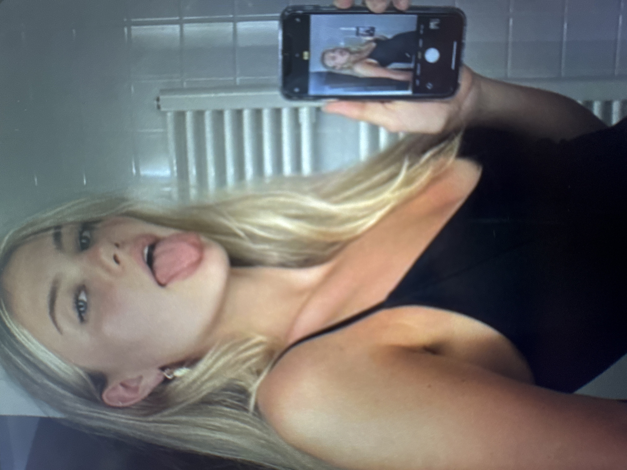 The image is oriented 90 degrees counter-clockwise, showing a close-up of a young woman taking a selfie. The woman has long, straight blonde hair that is spread around her, fair skin, and light-colored eyes. She is lying down, wearing a dark, possibly black, sleeveless top or dress. Her head is tilted, and she has a playful expression with her tongue slightly extended.
She is holding a dark-colored smartphone, likely an iPhone given the camera interface, above her with her right hand. The phone's screen clearly displays a live preview of the selfie she is taking, showing her in a similar pose with her tongue out. Visible on the phone screen are standard camera app controls, including a white circular capture button and other icons, though no specific text is legible.
In the background, a light-colored wall is visible, with a white radiator featuring vertical fins positioned behind and below her head. The lighting appears soft and even, suggesting an indoor setting. No discernible text is visible in the background or on her clothing.