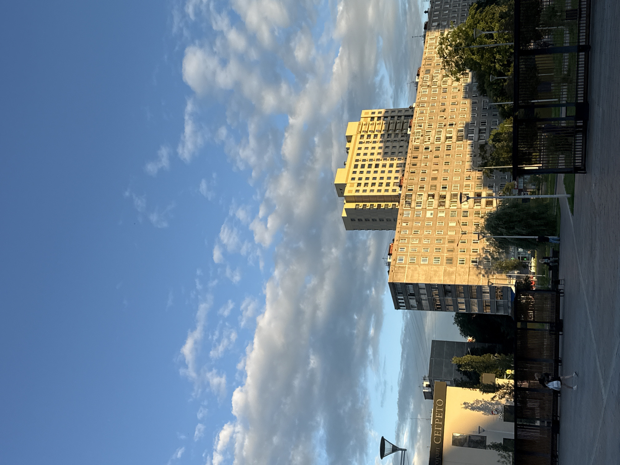 This media file depicts a street view in Moscow, Russia, with a focus on high-rise buildings under a partly cloudy sky. The scene is captured during daylight, likely late afternoon or early evening, given the warm, golden light illuminating the buildings and casting long shadows.
The primary subjects are the distinct apartment buildings. One is a tall, light-colored building with a facade that appears to be made of concrete, characterized by numerous windows. To its left, a slightly shorter, yellow-toned building with a more modern design is visible, also featuring many windows. Further in the background, another, darker high-rise building can be seen.
The foreground includes paved walkways and dark, slatted fencing, suggesting a public area or park adjacent to the buildings. Several streetlights are present, including one prominently featured in the lower portion of the frame. There are a few people in the scene, appearing as small figures in the distance, engaged in casual activities such as walking. The presence of trees adds a natural element to the urban landscape.
The sky is a vibrant blue, with dramatic, elongated clouds that are illuminated by the sun, creating a visually striking contrast.
A notable detail is the visible text on a building in the lower right quadrant of the image. The text reads "РЕСТОРАН СЕГРЕТО" (Restoran Segreto), indicating the presence of a restaurant with this name.
The overall impression is one of a typical urban environment in Moscow, bathed in the pleasant light of a late summer or spring day.