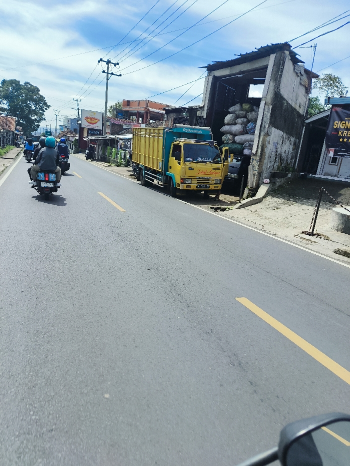 A yellow and green cargo truck is parked on the right side of a two-lane asphalt road in Ciwidey, Indonesia. There are three motorcycles traveling on the road. The truck is parked partially under a concrete structure. The structure appears to be partially collapsed and covered in debris. The truck is facing the camera and is partially obscured by the structure. There are a few buildings in the background with trees and power lines. The sky is blue and partly cloudy. There are three men riding the motorcycles. The camera is positioned on the back of one of the motorcycles, looking straight ahead toward the truck. The road is slightly curved, with a yellow line down the center. There is a yellow line on the right side of the road. The road is in good condition, with a few cracks. The scene was taken from a moving motorcycle.