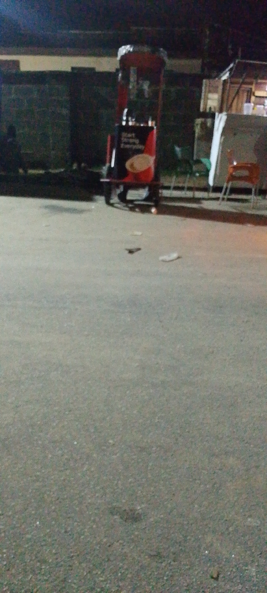 A night scene in Lokoja, Nigeria features a mobile food vending cart with a red frame and product advertisements on its sides. The cart is positioned on a grey asphalt surface in front of a block fence. A partially obscured figure sits near the fence. In the background are outdoor chairs and what appears to be a small structure or stall with white covering partially visible. The foreground is dominated by the asphalt surface with scattered small debris.