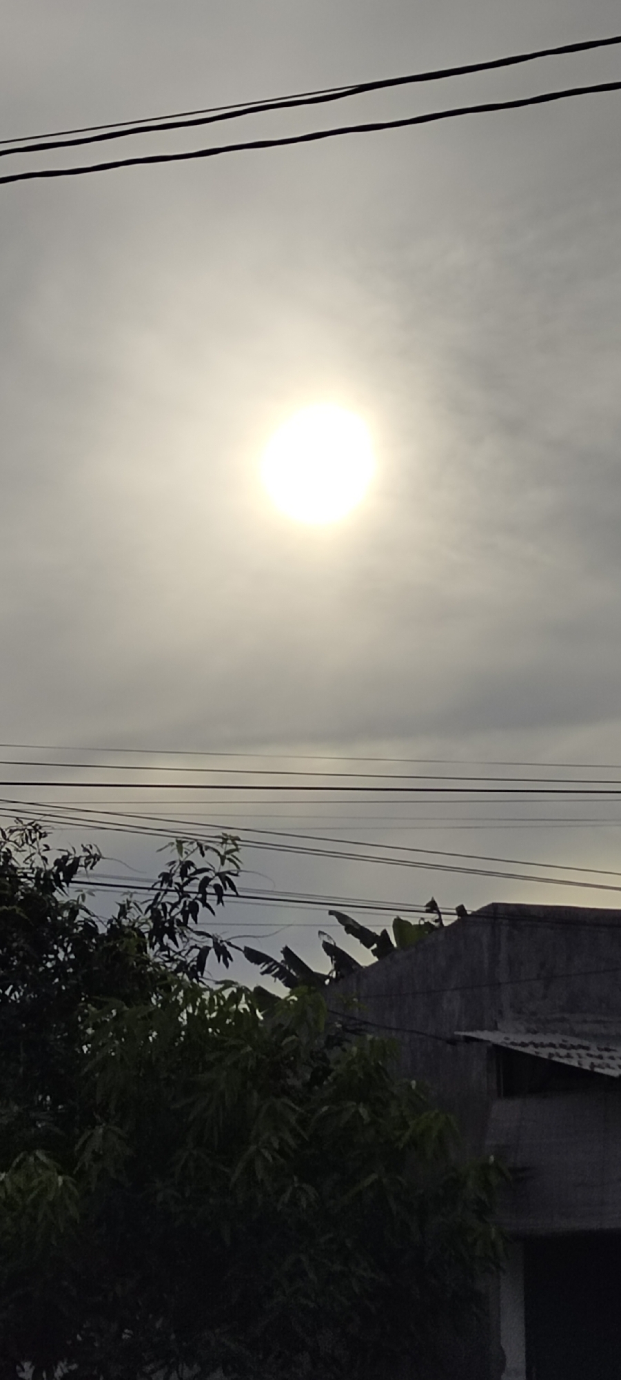 The image depicts a hazy sky with a bright sun obscured by clouds, taken in Lhokseumawe, Indonesia. In the foreground, there are trees and a concrete building with a corrugated roof. Electrical wires are visible crossing the sky. No people are visible.