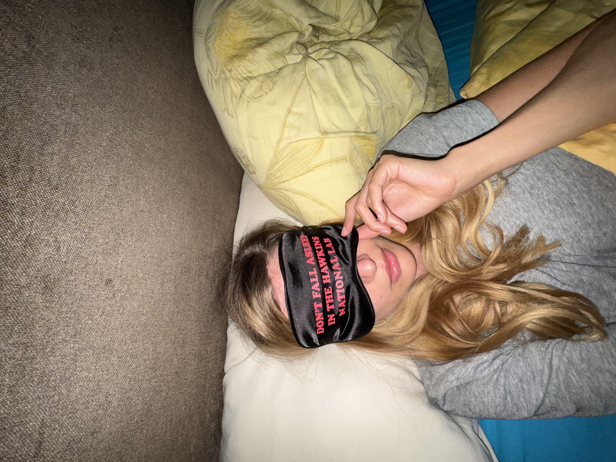 A blonde woman with a "DON'T FALL ASLEEP IN THE HAWKINS NATIONAL LAB" sleep mask lies on a pillow on a bed with a patterned yellow pillow and a gray shirt in Unknown, Unknown.