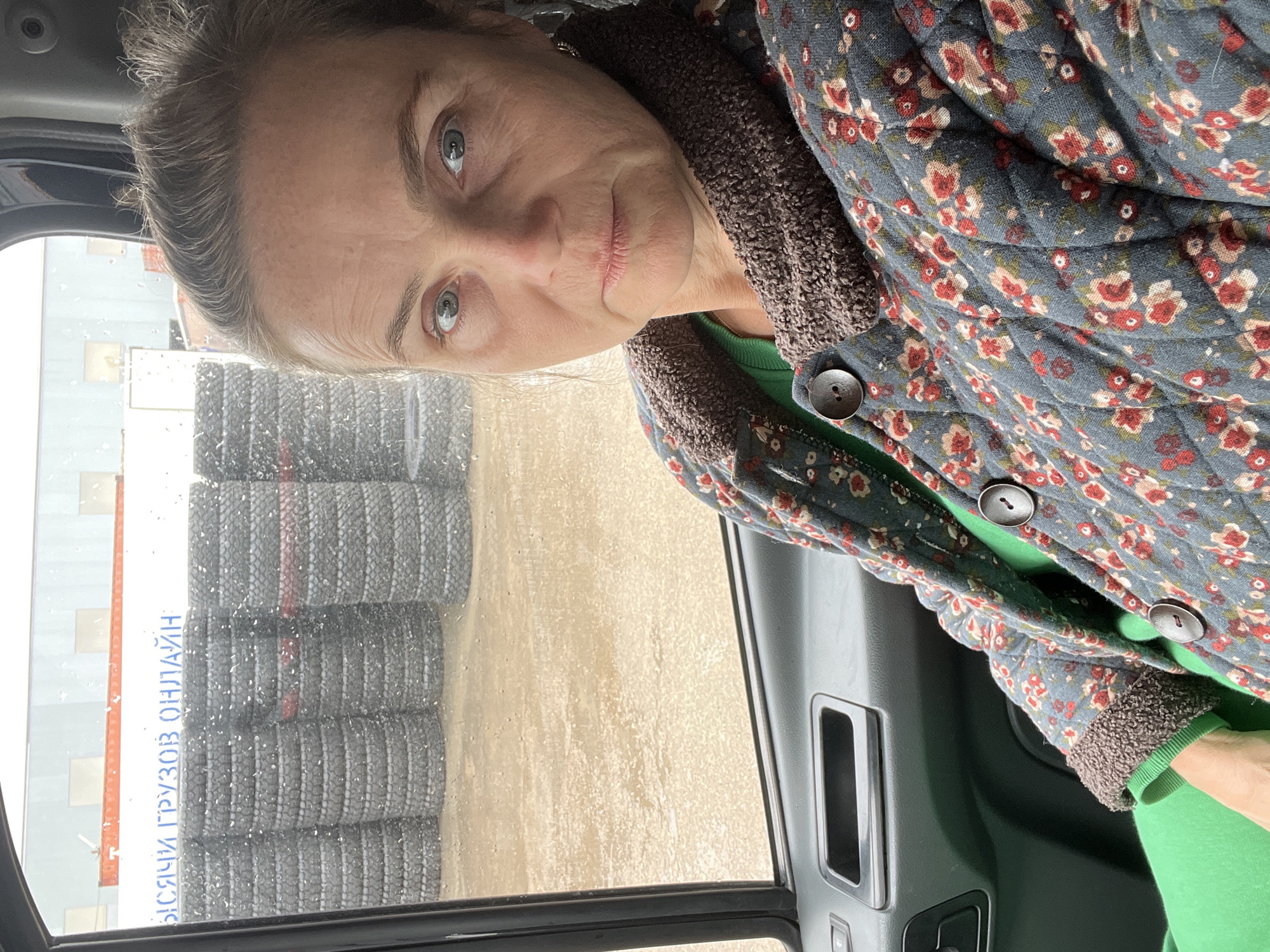 A woman is depicted from inside a vehicle, looking directly at the camera. She appears middle-aged with light-colored hair pulled back and light blue or grey eyes, exhibiting a neutral expression. She wears a bright green long-sleeved top under a dark grey or blue quilted jacket featuring a floral pattern of red, cream, and green flowers. The jacket has a textured brown collar and cuffs, with dark buttons visible on the front.
The interior of the vehicle, likely a car or truck, is visible on the right, showing dark panels and door features. Outside the window, which is speckled with raindrops, a wet and possibly muddy ground surface is present. Beyond the window, large stacks of dark tires, appearing to be truck tires, are visible. In the background, part of a building or structure can be seen.
The scene suggests a commercial or industrial setting, possibly a tire service center or a logistics yard. A sign behind the tires displays text in Cyrillic, which reads "ЫСЯЧИ ГРУЗОВ ОНЛАЙН," translating to "THOUSANDS OF TRUCKS ONLINE" or "THOUSANDS OF CARGO ONLINE." The image captures a daytime scene with evidence of recent precipitation. The location is Naberezhnye Chelny, Russia.