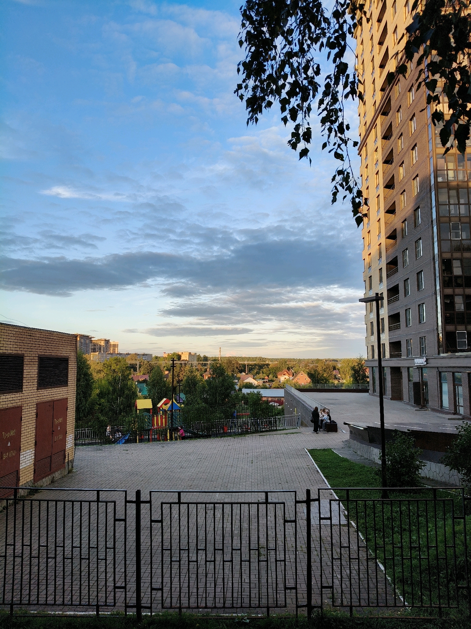 The image shows a multi-story apartment building in the city of Unknown, Unknown. The building has a facade of light brown brick and a section with dark glass windows. In the foreground, there is a black metal fence. Beyond the fence, there is a paved courtyard with a playground visible in the middle ground. A group of people are sitting on a bench near the playground. Further in the background, there are trees and other buildings, suggesting a suburban or residential area. The sky is partly cloudy. The time of day appears to be late afternoon or early evening, given the lighting.