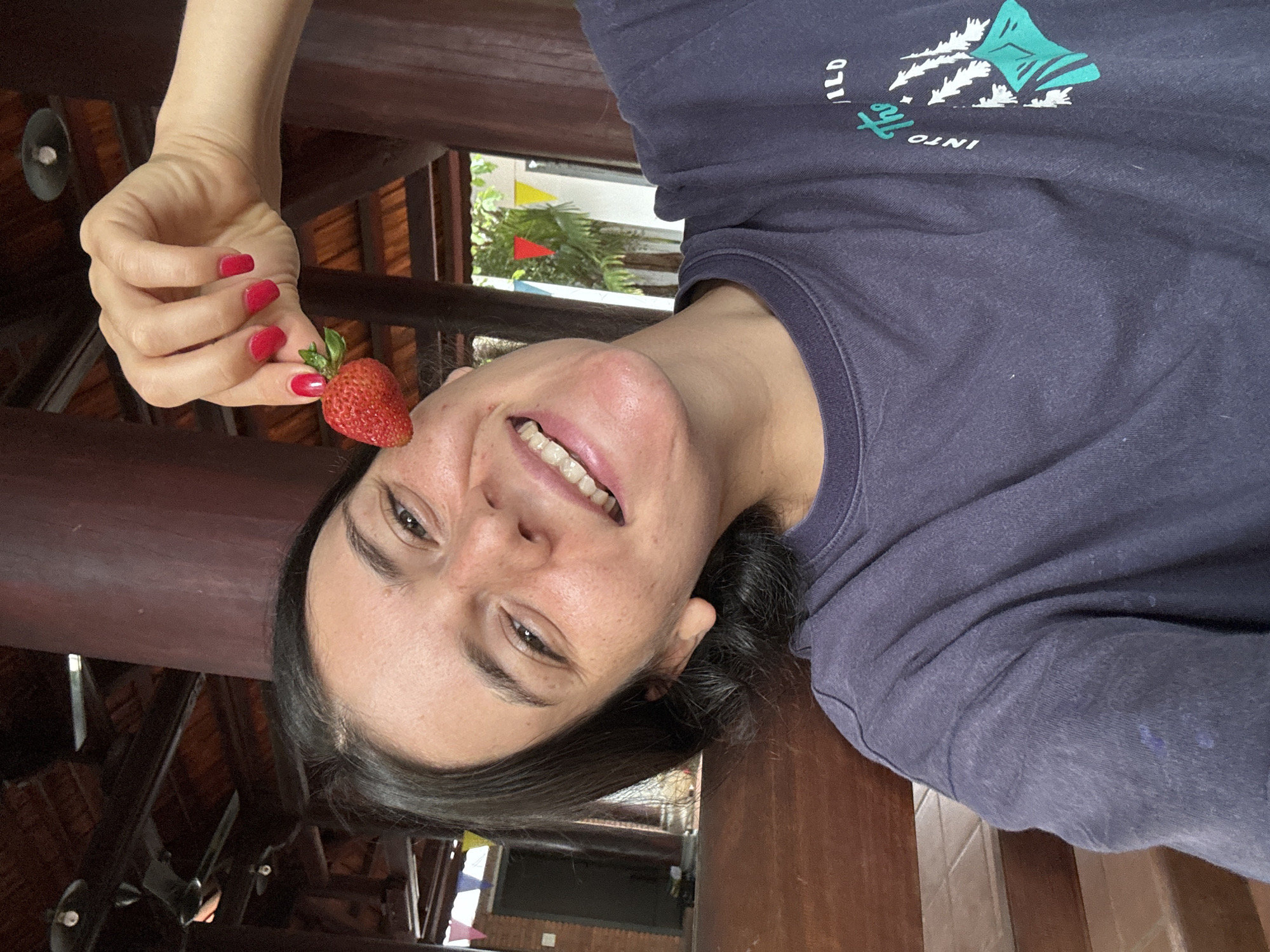 A young woman with dark hair and freckles is holding a strawberry up to her face in a selfie. She is wearing a dark navy t-shirt with a graphic design on the front that reads "Into the Wild". She has bright red nail polish on her fingernails. The background appears to be an outdoor covered area with wooden beams and a tiled floor, possibly a porch or pavilion. In the background, there are glimpses of tropical foliage and colorful triangular flags, suggesting a location in Mũi Né, Vietnam. The overall lighting is natural and appears to be daylight.