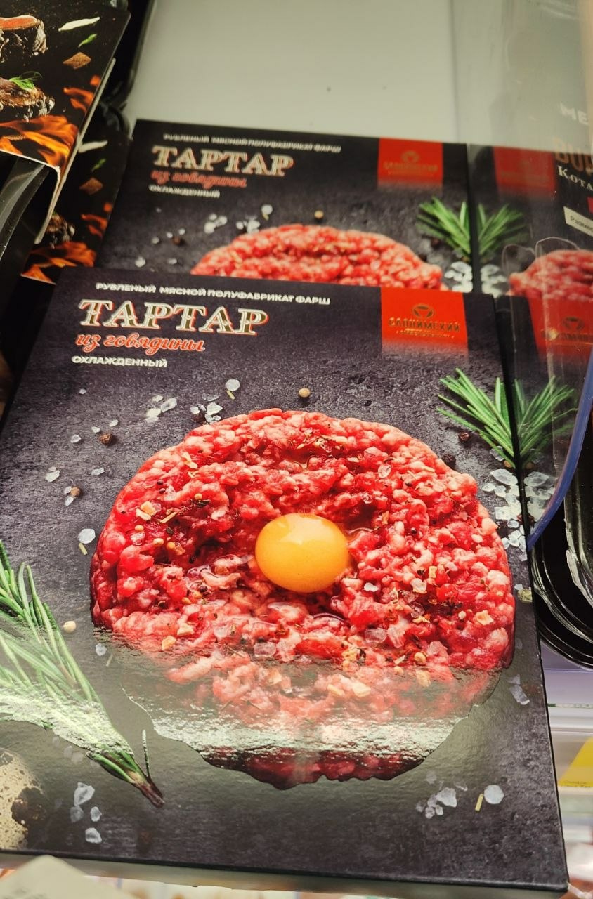 The image shows multiple packages of beef tartar displayed in what appears to be a store refrigerator. The foreground package is in focus, showing a close-up of the tartar with an egg yolk in the center, along with rosemary sprigs and scattered salt and pepper. The packaging is dark with red text in Russian. The name of the product, "TARTAR", is prominently displayed. Other similar packages are visible behind it. Based on the Cyrillic text on the packaging, the product is likely available in countries like Belarus. Although I cannot confirm a specific location in Minsk, Belarus without additional information, it is probable the picture was taken there as this is a place where Russian is one of the most popular languages used and beef tartar is available in stores.