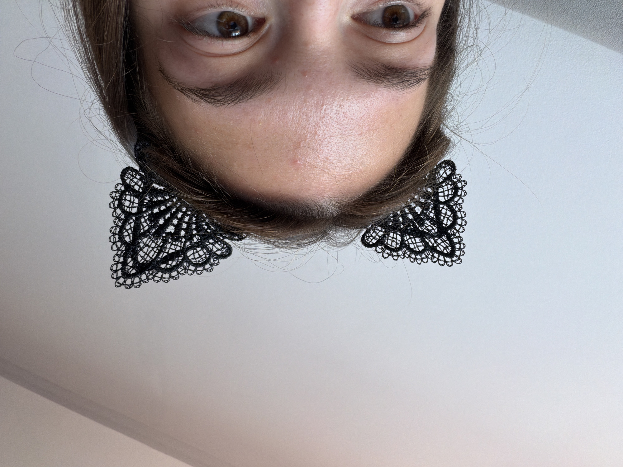 The image shows a close-up, upside-down view of a person's face, specifically focusing on their eyes and forehead. They have brown eyes and dark eyebrows. Their hair, which is brown and appears to be straight, frames their face. The person is wearing intricate black lace earrings that resemble bow ties. The background is a plain, light-colored surface, likely a wall or ceiling. The lighting suggests an indoor setting, possibly during the daytime given the brightness. No specific location cues or time of day are definitively discernible beyond it being indoors. The expression on the person's face is neutral. There is no visible text in the image.