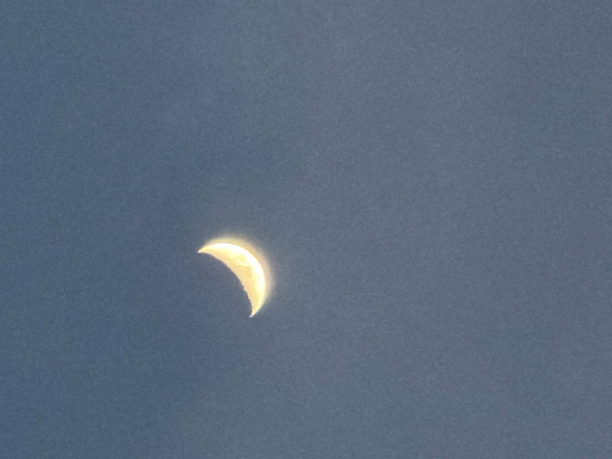 The image depicts a crescent moon in a dark blue sky. The moon is illuminated from the right side, with a bright yellow glow and subtle texture visible on its surface. The sky appears to be clear and cloudless. There are no other discernible objects or subjects in the frame. The image does not provide any visual cues that would allow for the identification of its location in Ko Samui District, Thailand.