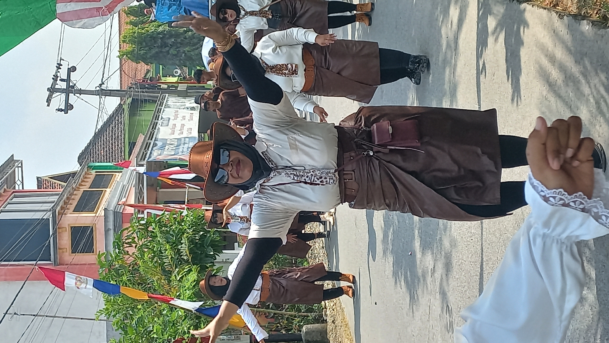 The image shows a street parade in Kedungmegarih, Indonesia. The foreground features a person's arm in a white shirt with lace cuffs, suggesting they are part of the parade, possibly filming or documenting. The central figure is a woman wearing a light-colored shirt, a patterned tie, dark sunglasses, a brown cowboy hat, and a brown wrap-around skirt. She has her arms outstretched, mid-dance. Behind her, other participants in similar attire, including brown hats and skirts, are also seen in motion. The street is lined with buildings, some with balconies, and decorated with flags. Power lines and trees are visible in the background. The overall scene depicts a festive cultural event.
