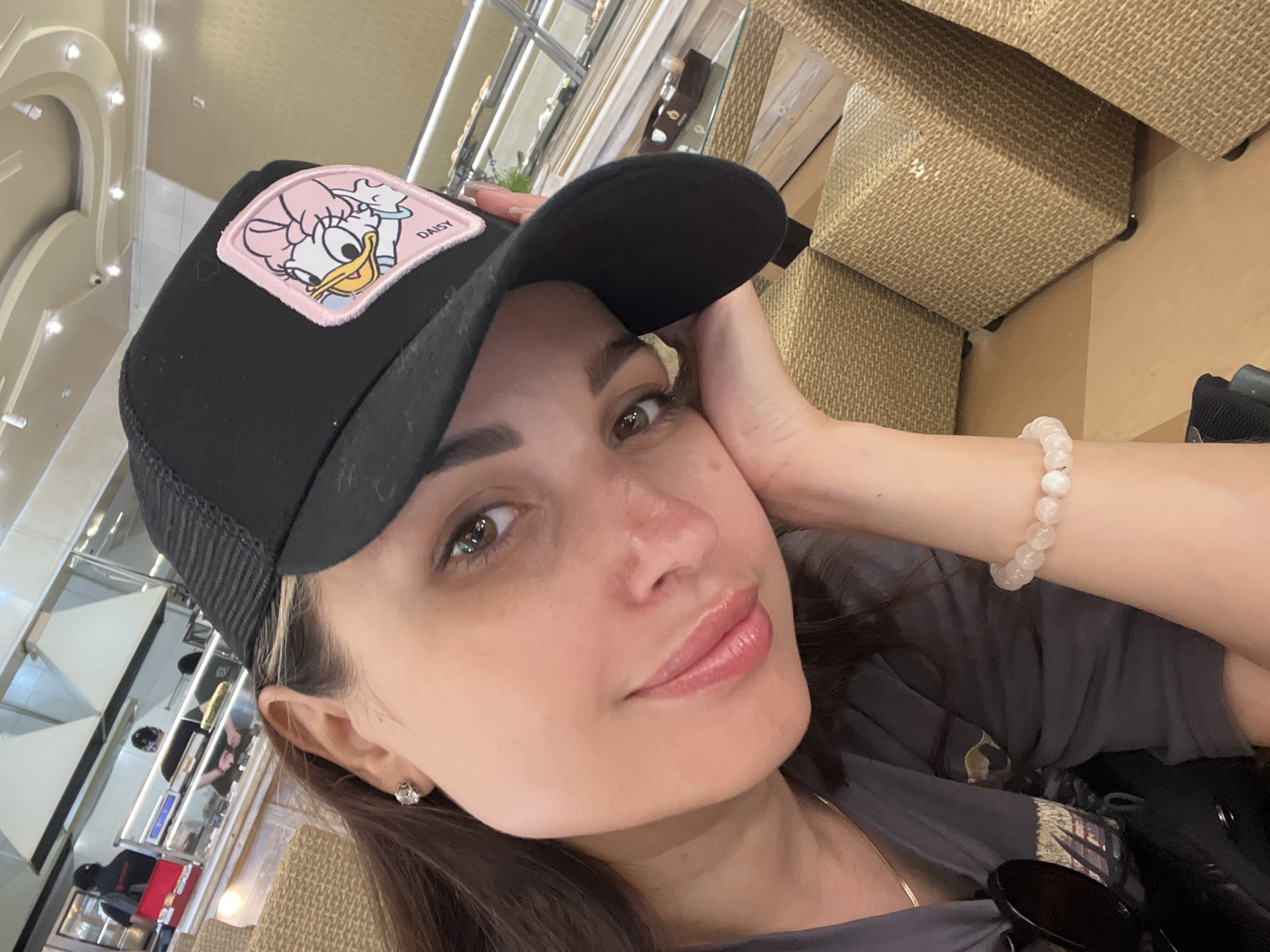 In an indoor setting, likely a cafe or restaurant with a food service counter in the background, a woman is captured in a close-up, selfie-style shot. She is wearing a black trucker hat with a patch of Daisy Duck on it, and a light-colored bracelet adorns her wrist. Her left arm is resting on her cheek, and her expression is relaxed and pleasant. The background shows stainless steel kitchen equipment and food display cases, with people in uniform working behind the counter. The overall lighting is bright, suggesting an overhead light source.