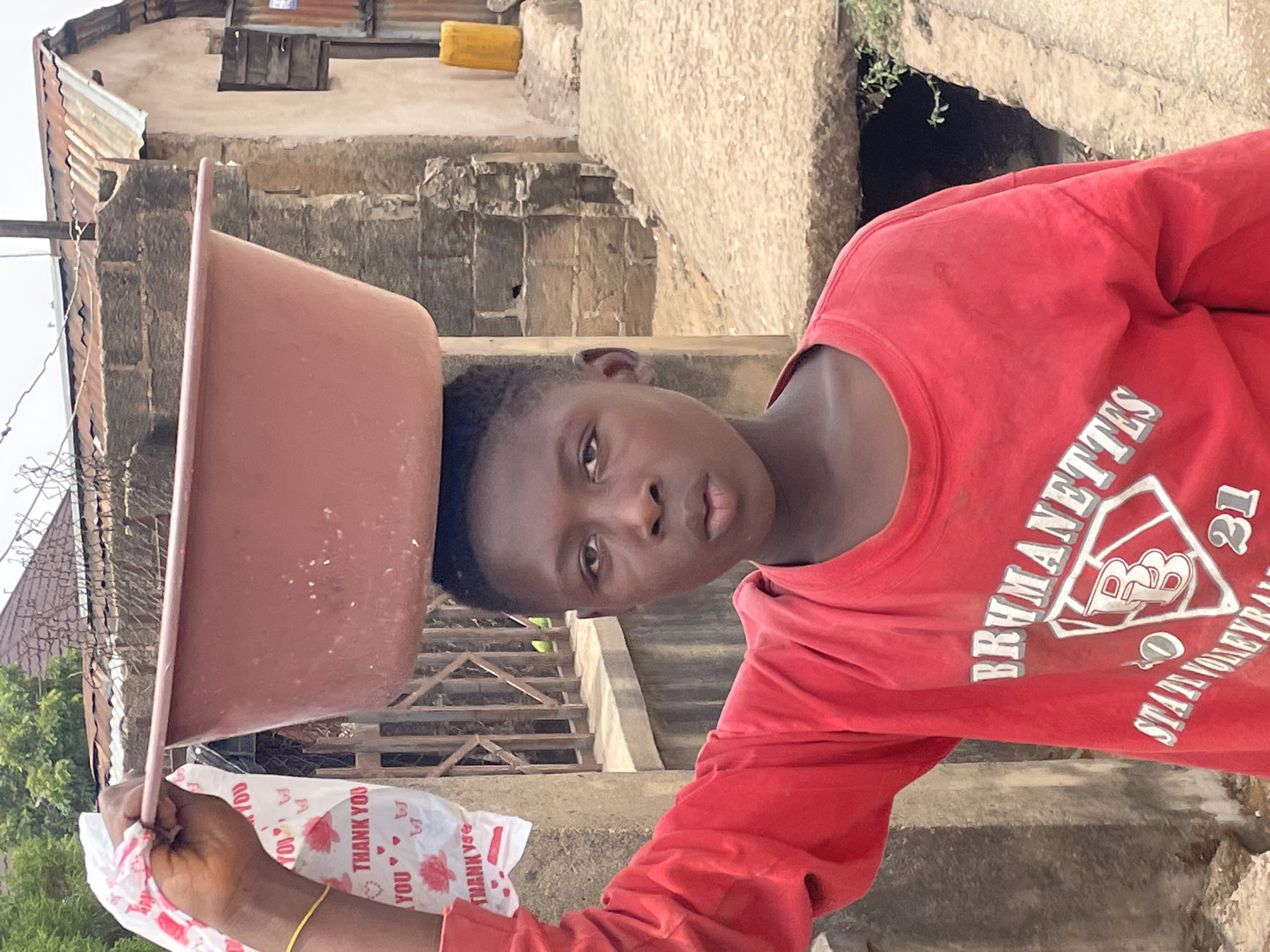 A young person, appearing to be a child or teenager, is depicted carrying a large, reddish-brown plastic basin on their head. They are also holding a white plastic bag with red text in their right hand, which reads "THANK YOU" repeated multiple times, interspersed with small heart symbols. The person looks directly at the camera with a neutral expression.
They are wearing a long-sleeved, vibrant red t-shirt. Visible text on the shirt includes "BRYMANETTES" (partially obscured), a logo featuring "BB" inside a diamond shape, and "STATE VOLLEYBALL 21" (also partially visible). A slender gold-colored bangle is worn on their right wrist.
The setting appears to be an outdoor, somewhat rustic area, likely a street or pathway in Jos, Nigeria. In the background, there are concrete structures, possibly walls or foundations, and what looks like corrugated metal roofing on a building. Some sparse vegetation is visible. The bright, even lighting suggests it is daytime, possibly morning or afternoon, with no harsh shadows. The act of carrying items on the head is a common mode of transport in many parts of Nigeria, aligning with the provided location context.