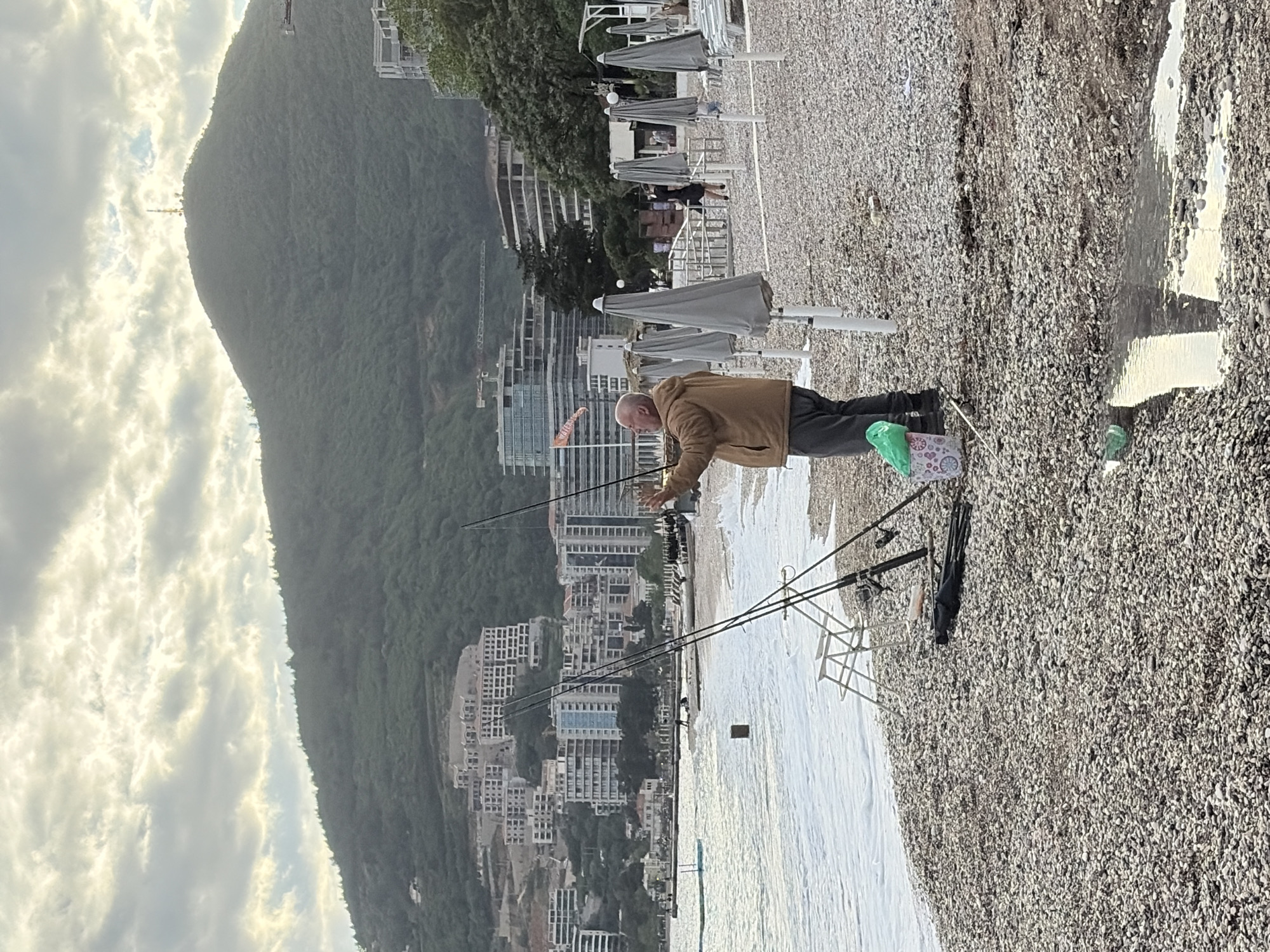 The image captures a man fishing on a pebble beach, likely in Budva, Montenegro, given the distinctive mountainous backdrop and coastal architecture. The man, dressed in a brown jacket and dark trousers, stands near the shoreline, actively holding a fishing rod. Several other fishing rods are set up in holders nearby. A bag and a water bottle sit near him. The backdrop includes modern buildings ascending up the hillside and lush greenery covering the mountain. Several beach umbrellas are visible. The sky is overcast. The beach is composed of small pebbles. Water washes onto the shore.