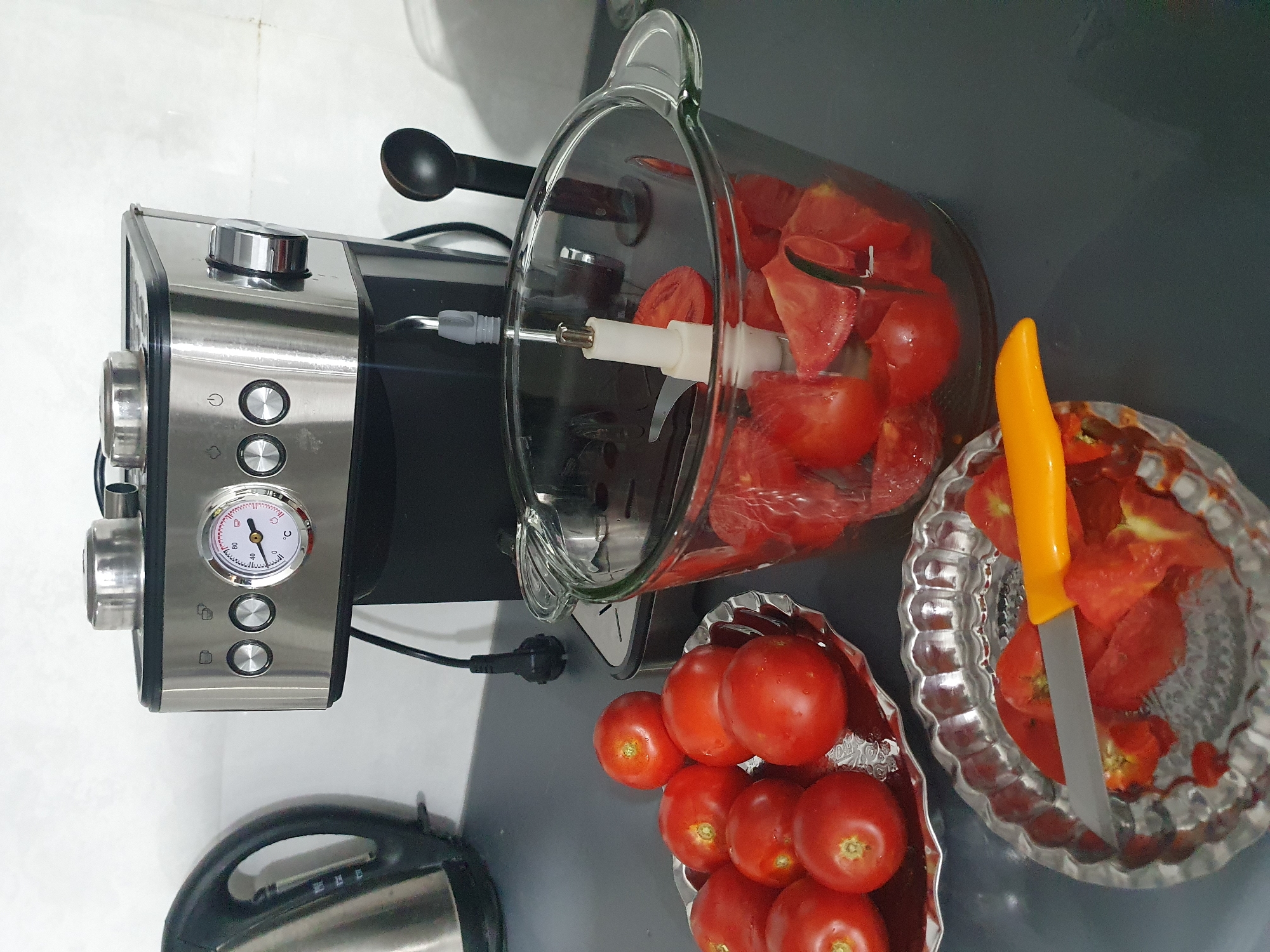 In a kitchen setting, a stainless steel coffee maker with a temperature gauge is positioned on a dark gray countertop. To the left of the coffee maker, a stainless steel electric kettle is partially visible. In the foreground, several ripe tomatoes are arranged on a decorative silver plate. Next to this plate, a clear glass bowl contains chopped tomatoes, suggesting preparation for a meal. A silver knife with an orange handle rests on another decorative silver plate, which also holds chopped tomatoes, indicating that the tomatoes are being processed. A black plastic spoon stands upright behind the glass bowl. The background consists of a plain white wall.