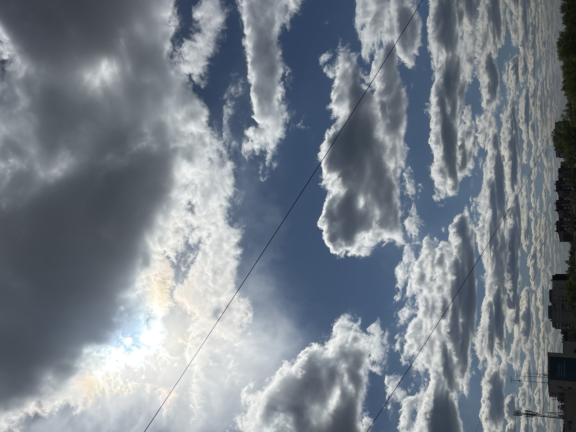 The image depicts a sky filled with cumulus clouds of varying sizes and densities. Some clouds are dark gray, indicating rain or storm conditions, while others are white and fluffy. Patches of blue sky are visible between the clouds. Overhead power lines diagonally cross the frame. The bottom right portion of the image reveals buildings and trees, indicating an urban setting. Based on the skyline and architectural style, this image is likely located in the city of Chaikovsky, Russia.