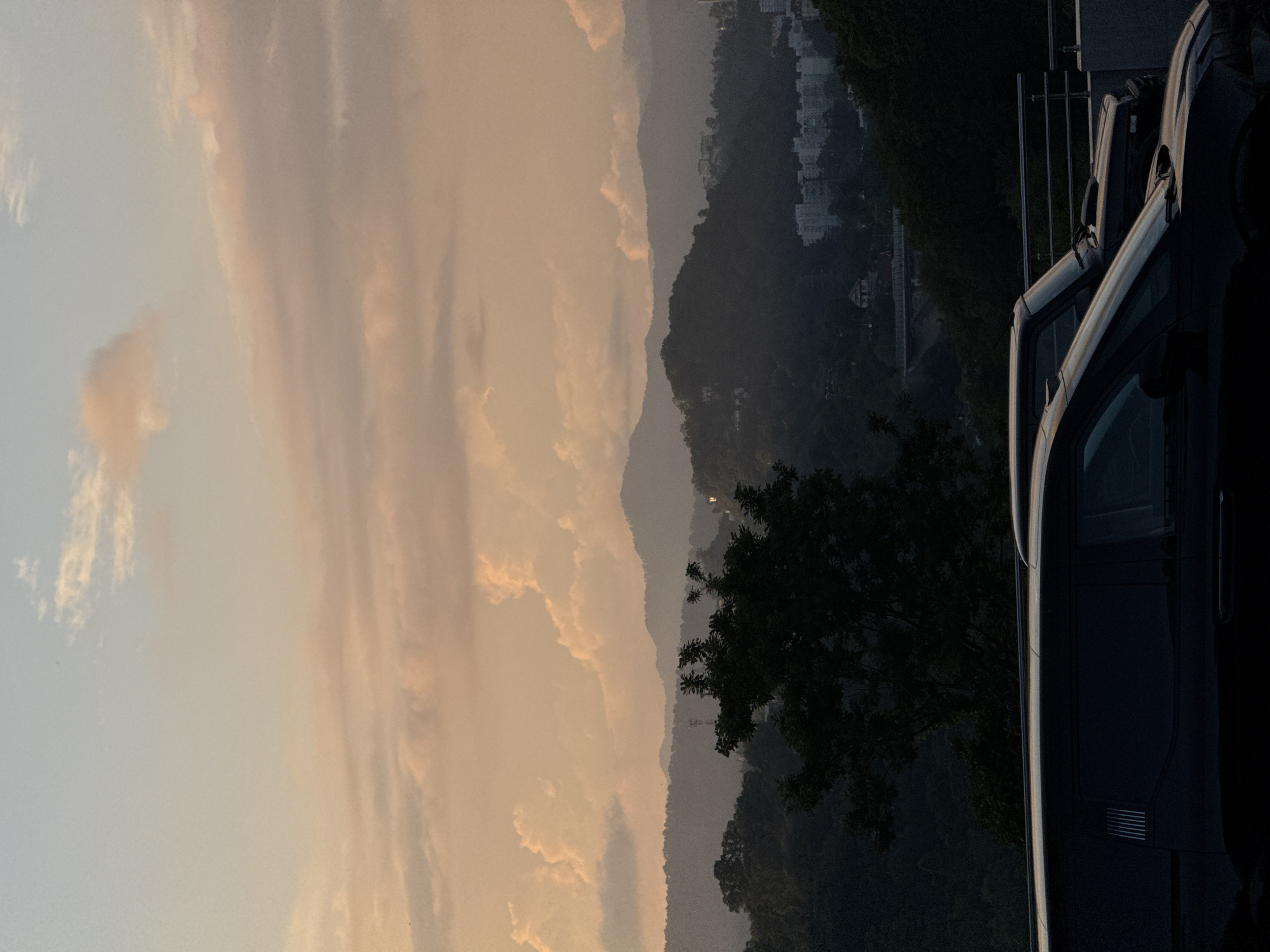 This image depicts a serene sunset scene in Sochi, Russia. In the foreground, two dark-colored vehicles are parked along a railing, their surfaces reflecting the soft, warm light of the setting sun. The railing itself is made of metal, with a smooth, silver finish. Beyond the vehicles, a lush green landscape unfolds, characterized by rolling hills covered in dense foliage. The hills are layered, with the furthest ones fading into a hazy, atmospheric perspective. A prominent tree with dark, spread-out branches stands in the middle ground, adding depth to the scene. In the distance, scattered residential buildings with white facades can be seen nestled amongst the hills, suggesting a settled urban or suburban area. The sky above is a canvas of soft colors, transitioning from a pale blue at the top to warm hues of orange and pink as it meets the horizon. Puffy, wispy clouds are scattered across the sky, catching the light and adding texture to the ethereal backdrop. The overall mood is tranquil and picturesque, capturing the natural beauty of Sochi at dusk.