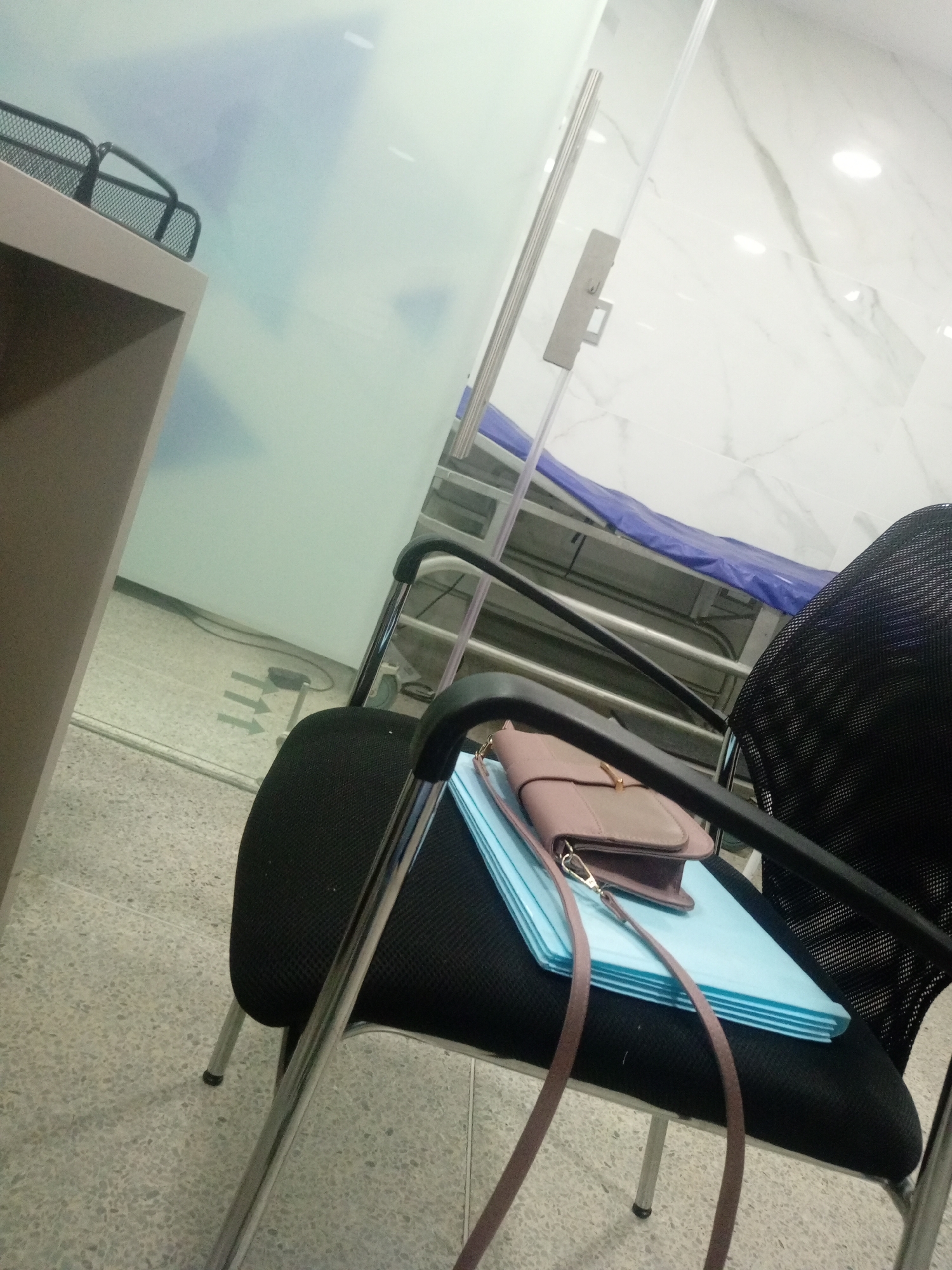 The image depicts an indoor scene, likely an office or waiting area, with a focus on a chair and its contents. A black mesh office chair is positioned in the foreground, with a stack of light blue folders on its seat. A small, light brown purse with a shoulder strap rests on top of the folders. The chair has chrome legs.
In the background, a glass partition separates the immediate area from another space. Beyond the partition, a wall with a marble-like pattern is visible, illuminated by overhead lights. A blue tarp-covered object, possibly a piece of medical equipment or furniture, is partially visible in the far background. To the left of the chair, a portion of a light brown desk or counter is visible, topped with a black wire basket. The floor is tiled with a speckled pattern.
The image does not contain any people and provides no explicit information about the location within Perímetro Urbano Cúcuta, Colombia. The overall atmosphere is neutral and functional.
