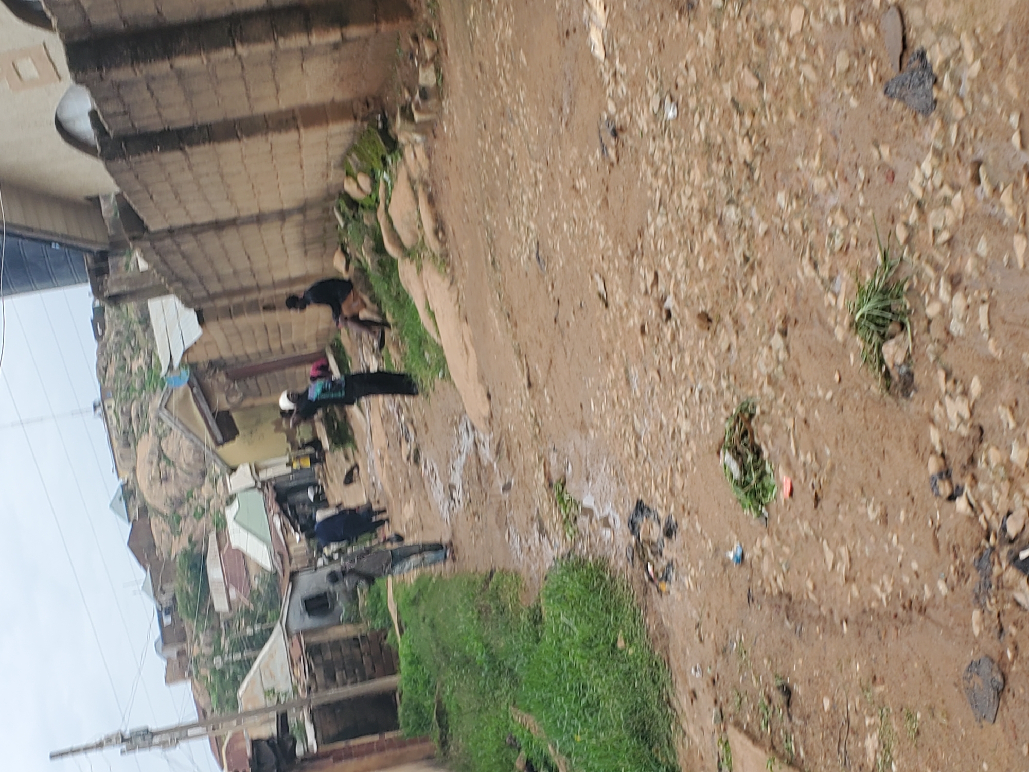 The image depicts a dirt road in Jos, Nigeria, with a slight incline leading towards a rocky hillside covered in scattered buildings. The road is muddy and appears to have puddles, suggesting recent rainfall. There are several individuals present: one person walks away from the camera down the road, another sits on rocks to the right, and two others are further down the road, with one appearing to be a child. Buildings made of brick, corrugated metal, and concrete line the sides of the road, some with visible roofs and others appearing partially constructed. A tall brick wall separates the road from some of the structures on the right. Green grass grows along the left side of the road, and scattered rocks and debris are visible throughout the scene. The sky is overcast.