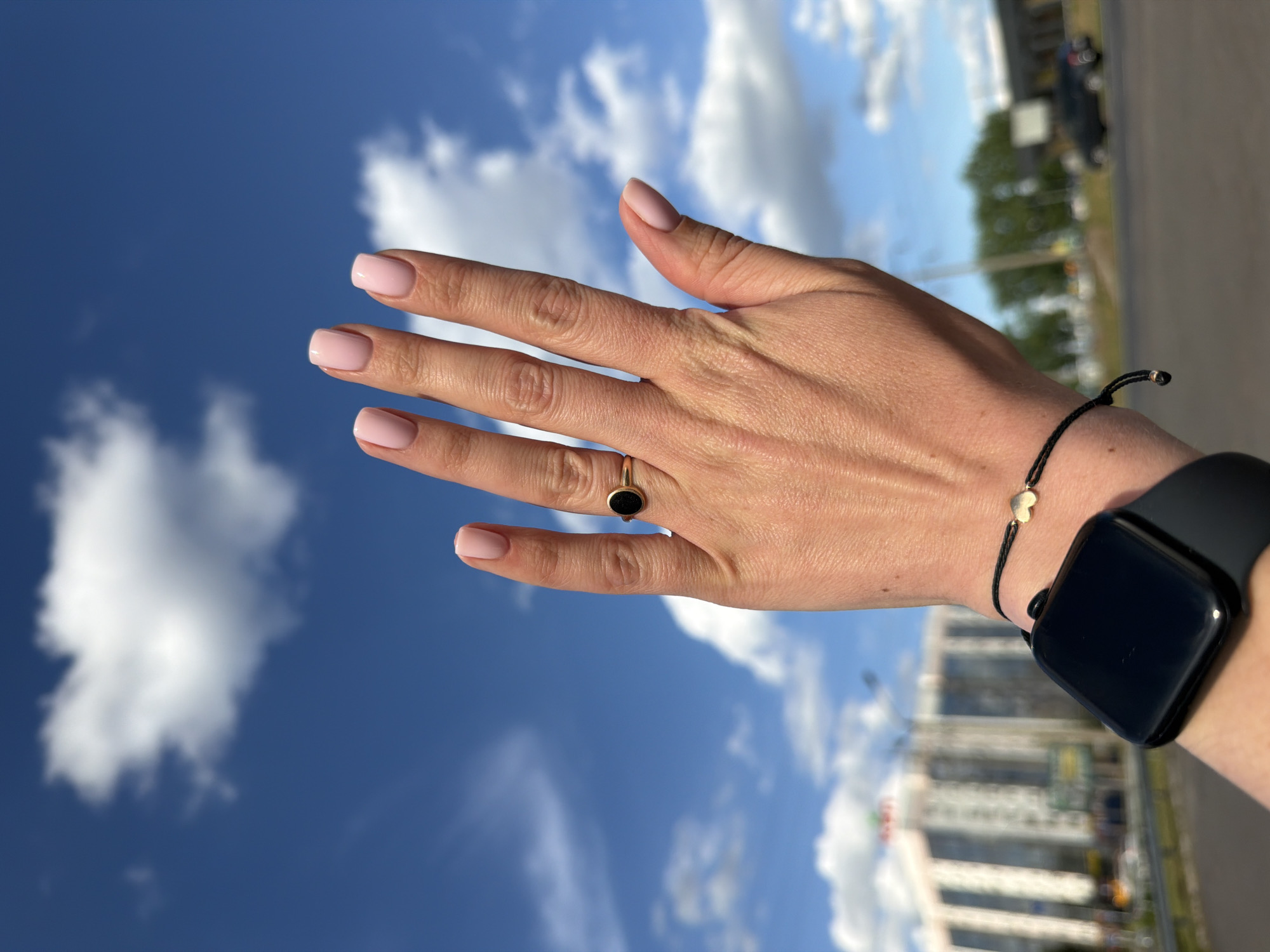 A person's hand is raised against a blue sky with white clouds. The hand has light pink nail polish. A gold ring with a black oval stone is on the ring finger. A black smartwatch and a black string bracelet with a gold heart charm are on the wrist. In the background, a building and trees are visible, along with a road and a car. The location is Unknown, Unknown.