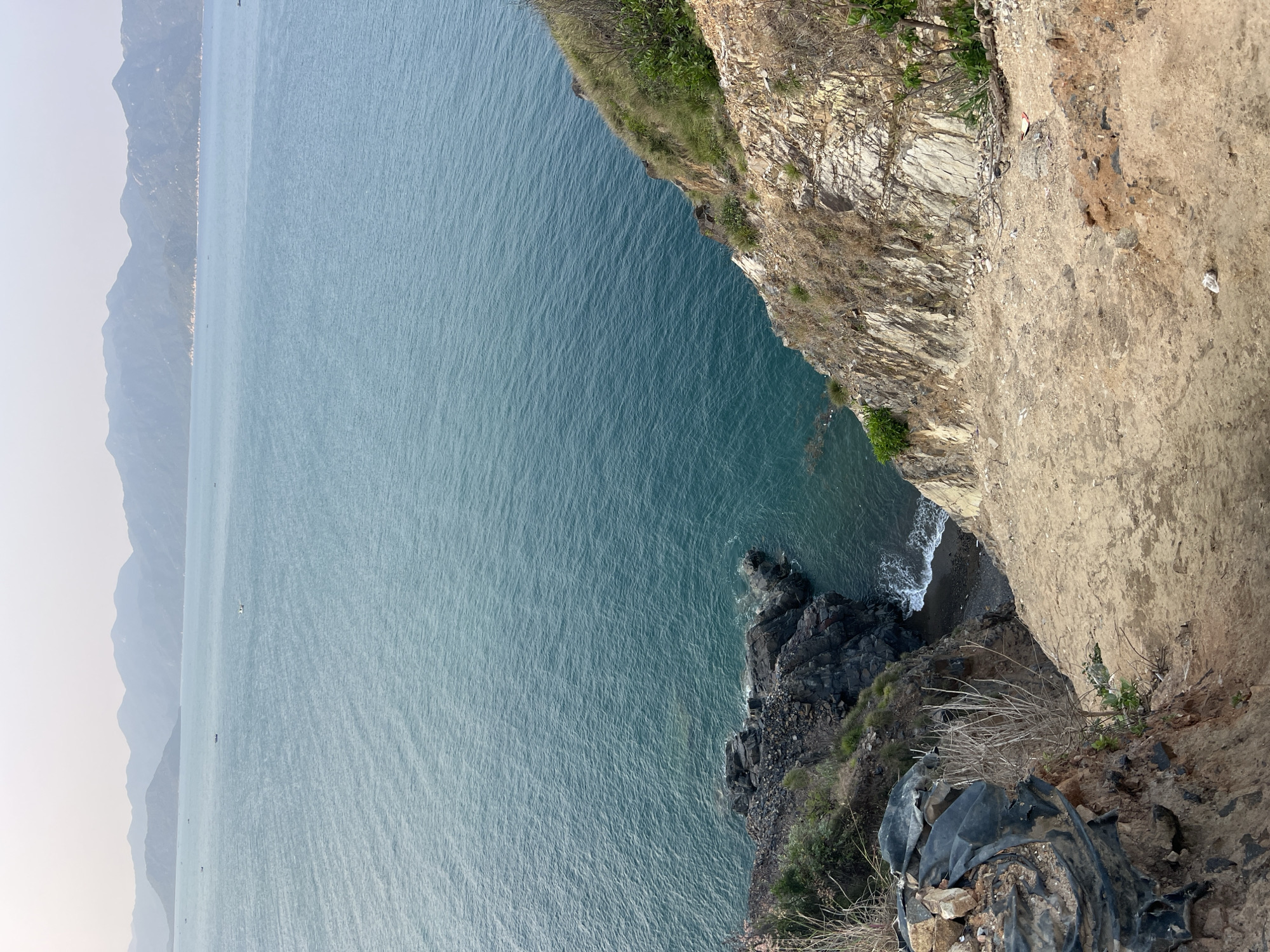 A view from atop a cliff in Bac Natural Heritage Area Trang, Vietnam, showcases a body of water to the left. The cliffside is characterized by stratified rock formations and earthy tones, with sparse vegetation clinging to the edges. Below, waves gently lap against a rocky beach. A black tarpaulin with debris is at the bottom of the frame. In the distance, the outlines of mountains fade into a hazy horizon. The water appears calm, its surface reflecting the sky.