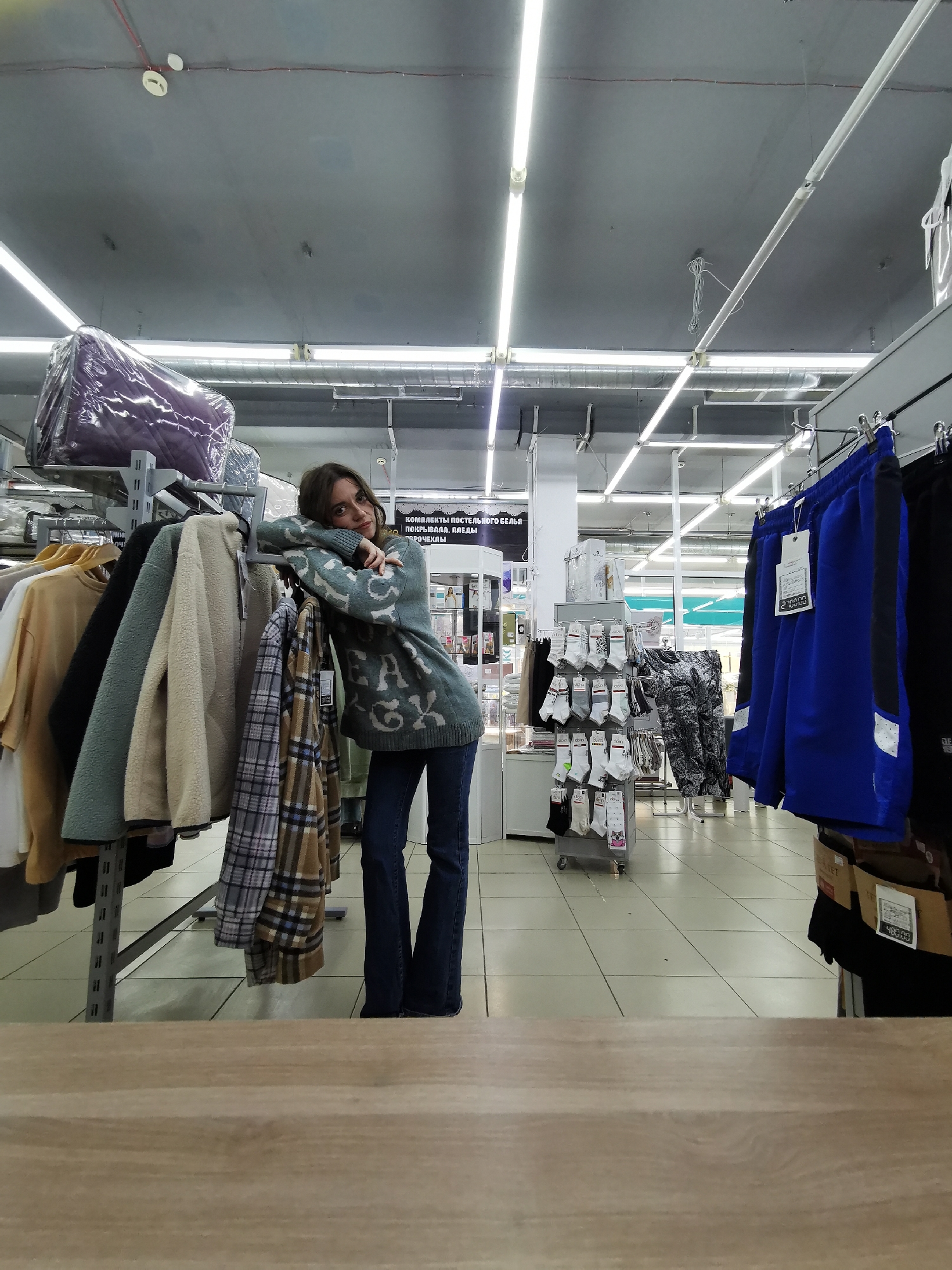A young woman is posing in what appears to be the clothing section of a retail store. She is positioned slightly to the left of the center, leaning casually on a clothing rack filled with garments. She has long, dark hair and is looking directly at the camera with a calm expression. She is wearing a greyish-blue knit sweater with a light-colored pattern that appears to spell out "GREY FOX" or similar text, and dark blue flared jeans.
The clothing rack she is leaning on displays various items including a beige fleece jacket, a teal/mint-colored fleece jacket, a plaid long-sleeved shirt (brown, blue, and white), and a yellow/tan t-shirt. To the right, another rack holds blue athletic shorts with white accents, one bearing a price tag of "2 700.00". Further in the background, a display stand showcases various white and colored socks. The store has a utilitarian aesthetic with a grey ceiling, visible industrial lighting fixtures (fluorescent tubes), red and white wiring, and a light-colored tiled floor. A wooden counter or surface is visible in the immediate foreground, suggesting the photo was taken from above it.
On a white sign in the background, text in what appears to be Russian reads: "КОМПЛЕКТЫ ПОСТЕЛЬНОГО БЕЛЯ, ПОКРЫВАЛА, ПЛЕДЫ, ПОДУШКИ" (Bed linen sets, bedspreads, throws, pillows). Another price of "480.00" is visible on a box or item near the blue shorts. The overall setting suggests an indoor retail environment during operating hours, given the bright lighting.