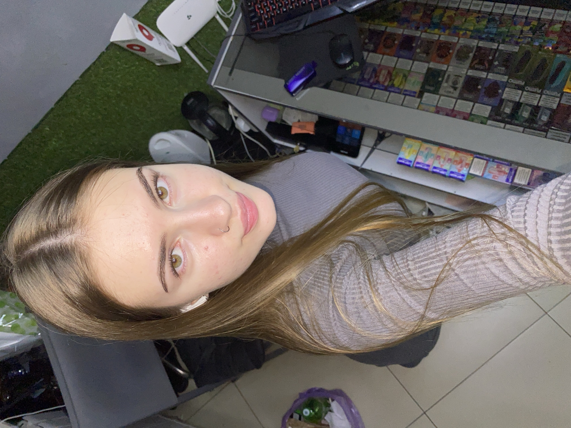 The image shows a young woman with long brown hair, wearing a grey turtleneck sweater. She has light skin with some freckles and blemishes, and her eyes are light brown. She is wearing white earbuds and a nose ring. The woman is positioned in what appears to be a shop or store, with shelves displaying various items. In the background, there is a grey desk, artificial turf, and electronic devices such as a computer monitor and keyboard. The overall lighting suggests an indoor environment. The location is identified as Dyakovo, Russia.