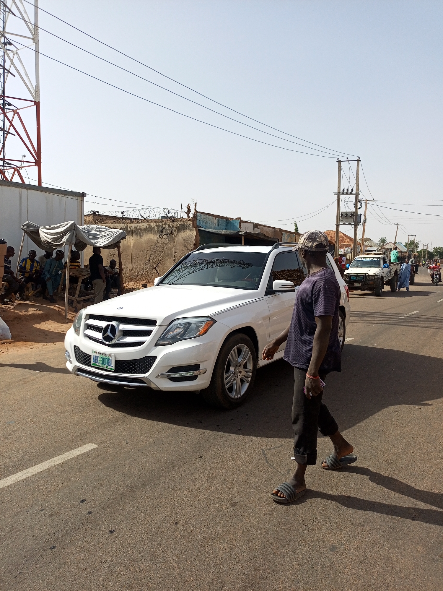 The scene is a street in Tempure, Nigeria. A white Mercedes-Benz SUV with license plate "ABC-300BL" is parked on the left side of the road. A man is walking in front of the car, toward the right side of the frame. He is wearing a patterned cap, a dark shirt, dark pants, and sandals. On the left side of the road, some men are sitting and standing under a shaded area near a building. In the background, other vehicles are visible on the road, including a white pickup truck. An electrical utility pole is visible in the midground. The sky is clear and the light suggests it is daytime. A metal tower is visible in the background on the left.