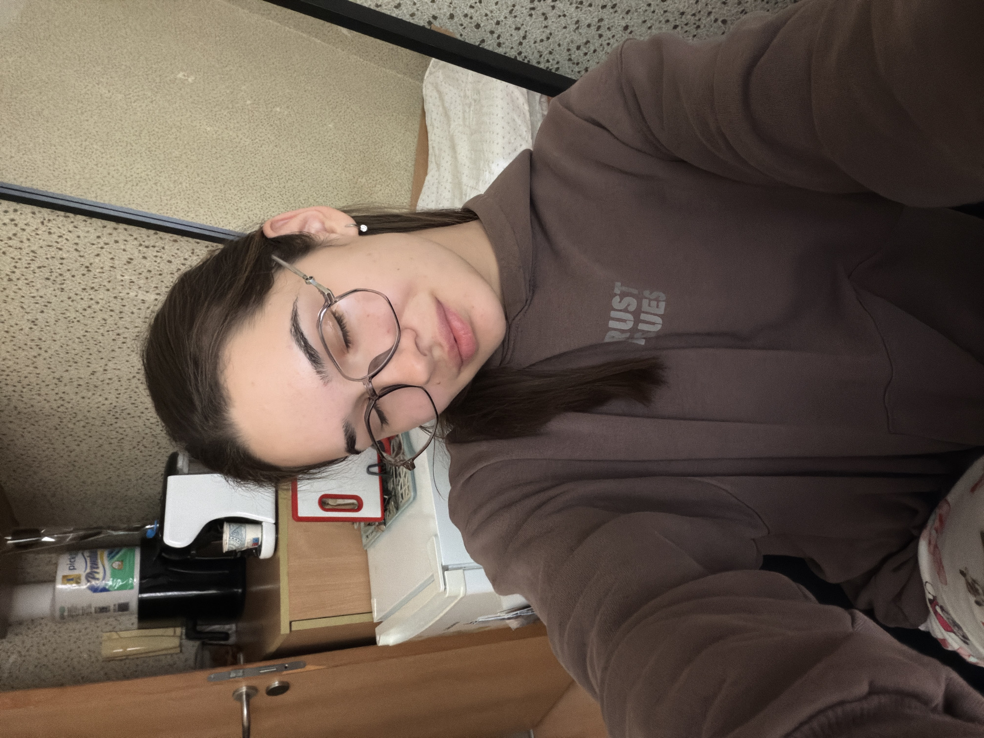 This media file is a close-up, rotated selfie featuring a young woman in an indoor setting, likely in Sofia, Bulgaria. The image is rotated 90 degrees clockwise.
The subject is a young woman with dark brown hair, wearing glasses with thin, possibly metallic frames. Her eyes are closed or partially closed, and her lips are slightly parted, suggesting a moment of relaxation or rest. She wears a brown or muted taupe-colored hoodie, on which the text "RUST RUES" is visible. A small, shiny earring is visible in her left ear.
The background on the left side of the image (which would be below her head if correctly oriented) shows several objects on what appears to be a desk or shelf. These include a white and green bottle with the partially visible label "pickles Premium," a black and white device resembling a stapler or label maker containing a rolled item, sections of wooden furniture, a white printer or small appliance, and a red and white item that could be a cutting board. On the right side of the image (above her body), a white patterned pillow and another patterned fabric object are visible. The wall or ceiling in the background is light-colored and has a textured, speckled appearance. A dark, thin bar or frame runs across the top part of the background. The lighting is soft, not indicating a specific time of day.