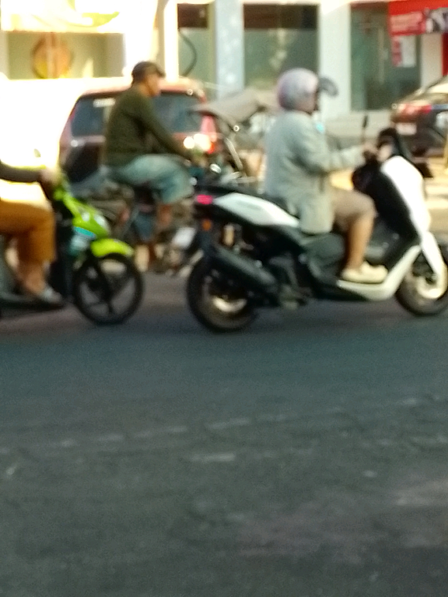 In Jember, Indonesia, two individuals are riding scooters on a street. The person on the left, wearing a dark long-sleeved shirt and light blue pants, is on a green scooter. The person on the right, wearing a light-colored jacket and tan pants, is on a white scooter and has a purple helmet. Both are in motion, with the scooters' wheels visible. The background shows buildings, suggesting an urban environment. The overall image is slightly blurred, indicating movement or a focus on the subjects rather than sharp detail.