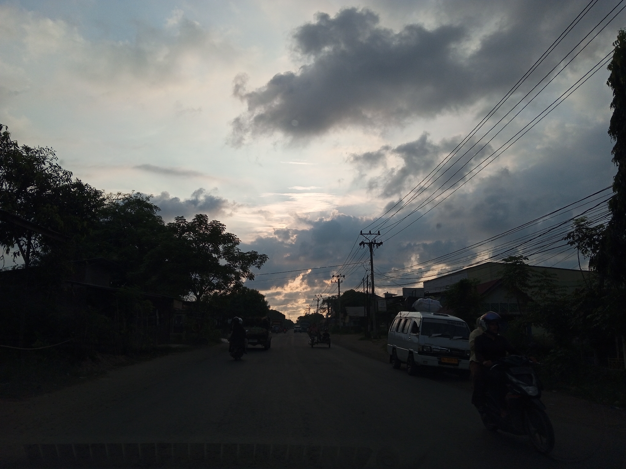 The image depicts a street scene in Palohlada, Indonesia, under a cloudy sky with shafts of sunlight breaking through. A paved road stretches into the distance, flanked by trees and buildings. Several vehicles are visible, including a white van, motorcycles, and what appears to be a three-wheeled vehicle. Power lines are strung across the scene, supported by utility poles. The overall impression is that of a rural or semi-urban area during the late afternoon or early evening.