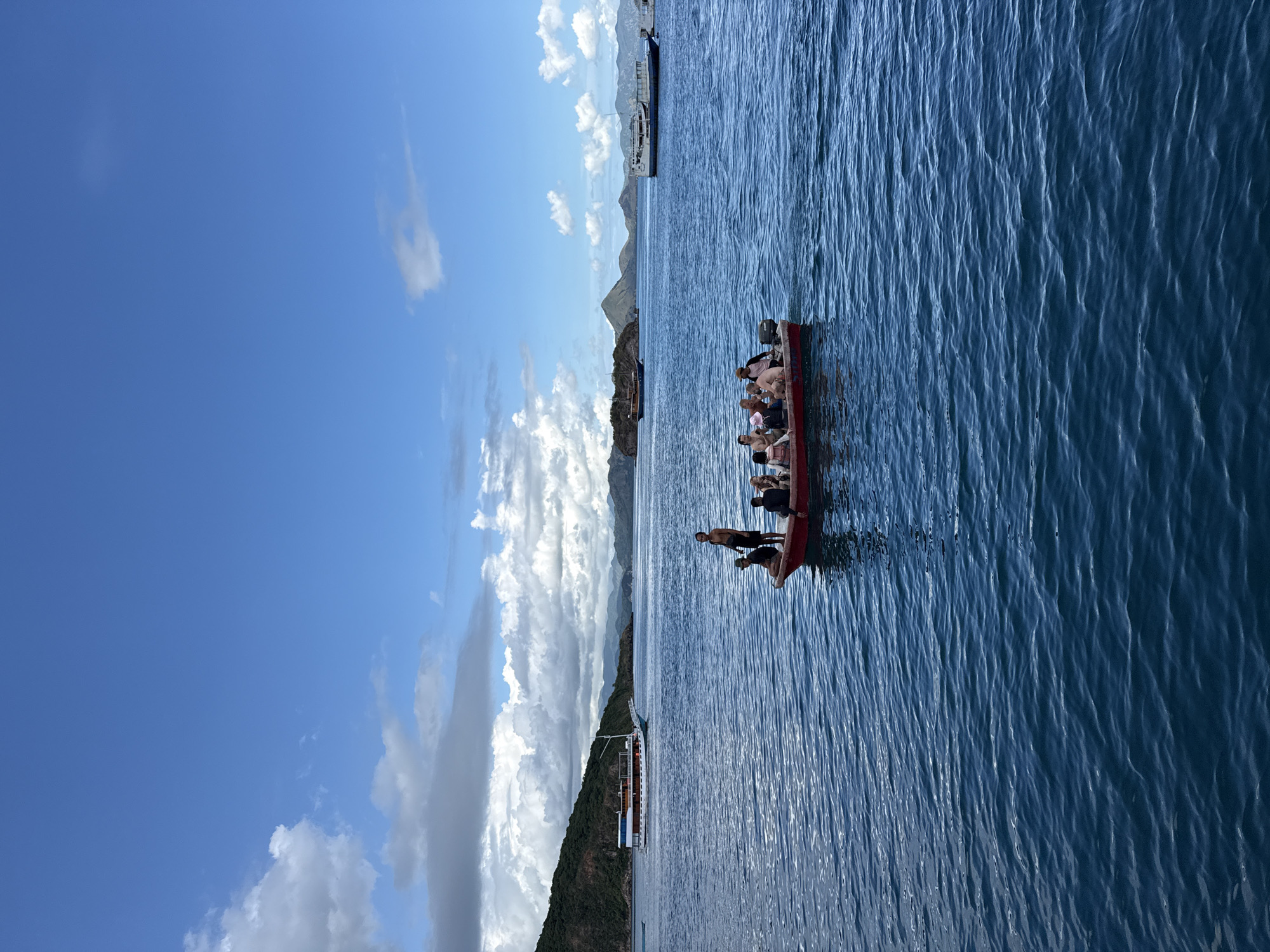 The image shows a boat carrying passengers on the ocean in Pasir Putih, Indonesia. In the background, there is land with mountains. The sky is blue with white clouds. The ocean water is blue and rippled.