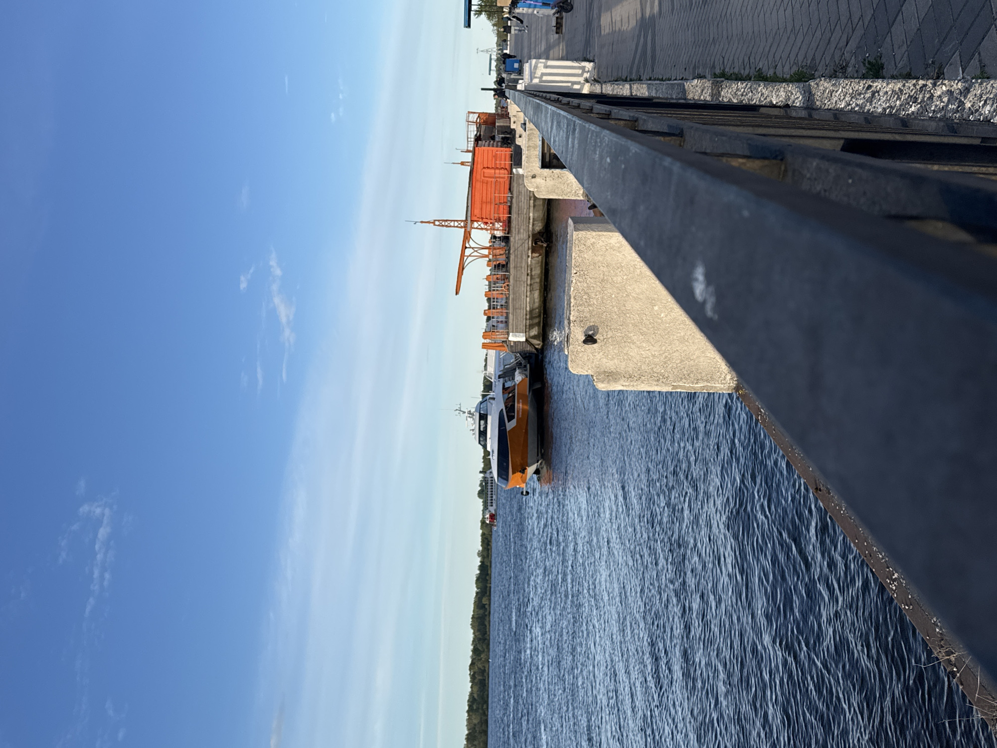 The image shows a view from a bridge or elevated walkway overlooking a body of water, likely a river, in what appears to be an urban or industrial setting. A modern passenger boat, characterized by its orange and white paint scheme, is docked alongside a concrete pier or embankment. On the pier, there is an orange crane. The setting is near the city of Nizhny Novgorod, Russia, in the foreground of the image is a metal railing. The sky is blue with some wispy clouds. Along the top of the image is a concrete walkway and sidewalk.