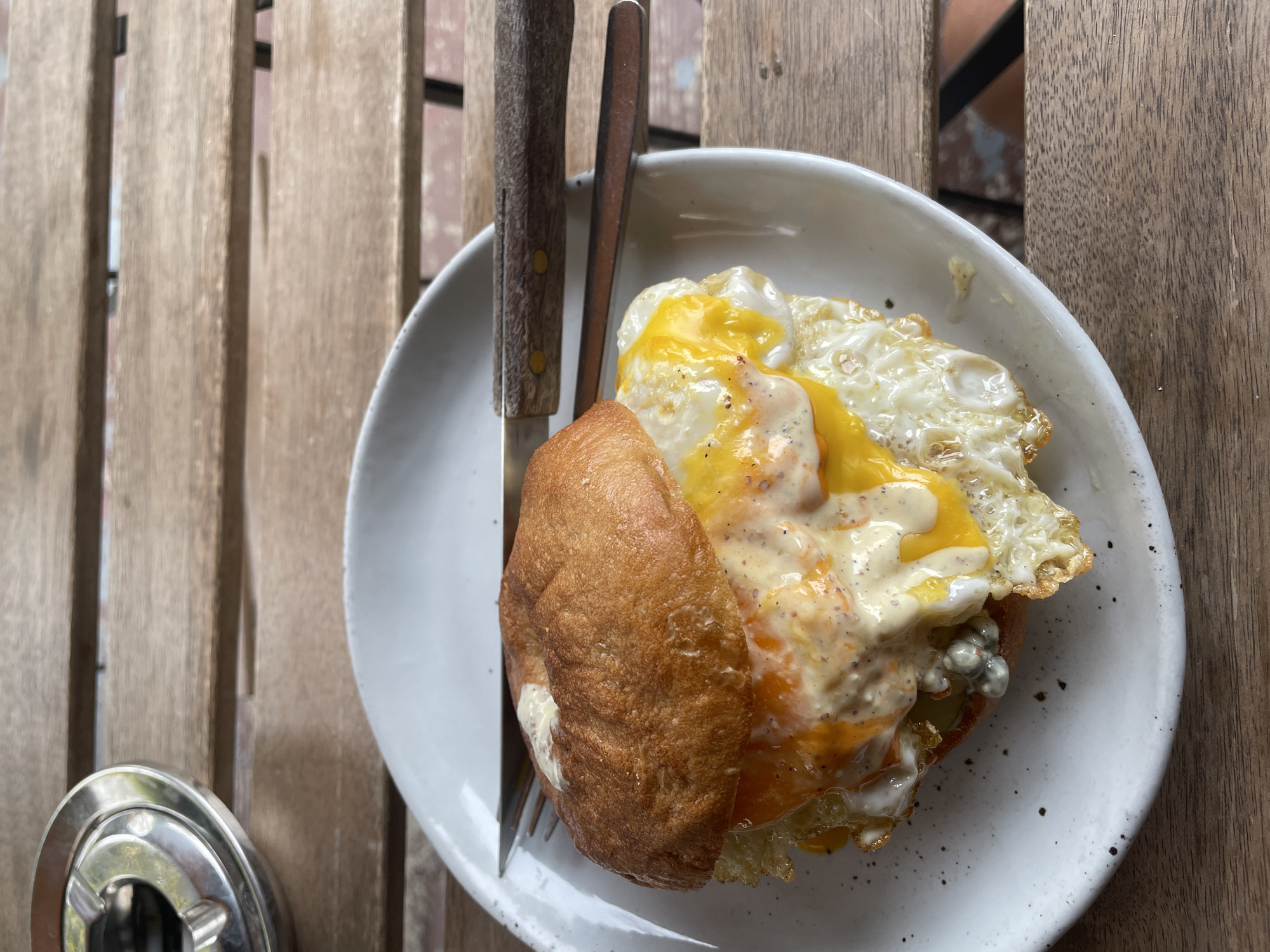 An overhead, close-up shot displays a hearty meal served on a white ceramic plate, resting on a wooden slatted table. The main dish appears to be a breakfast or brunch sandwich, featuring a glossy, golden-brown bun. Two fried eggs with vibrant yellow yolks and solidified whites, generously covered in a creamy, speckled sauce (likely a mayonnaise or hollandaise with pepper), top the bun. Beneath the eggs, some green or greyish ingredients are visible, possibly pickled vegetables or a relish.
To the left of the plate, a knife and fork with rustic wooden handles are neatly placed. The table is constructed from light brown wooden slats, showing natural grain and texture. In the bottom left corner, a portion of a round, metallic ashtray is visible. The lighting suggests it is daytime, suitable for a breakfast or brunch setting. The overall scene conveys a casual dining experience, possibly at an outdoor cafe or restaurant in Chiang Mai City Municipality, Thailand. No people are visible, and there is no discernible text in the image.