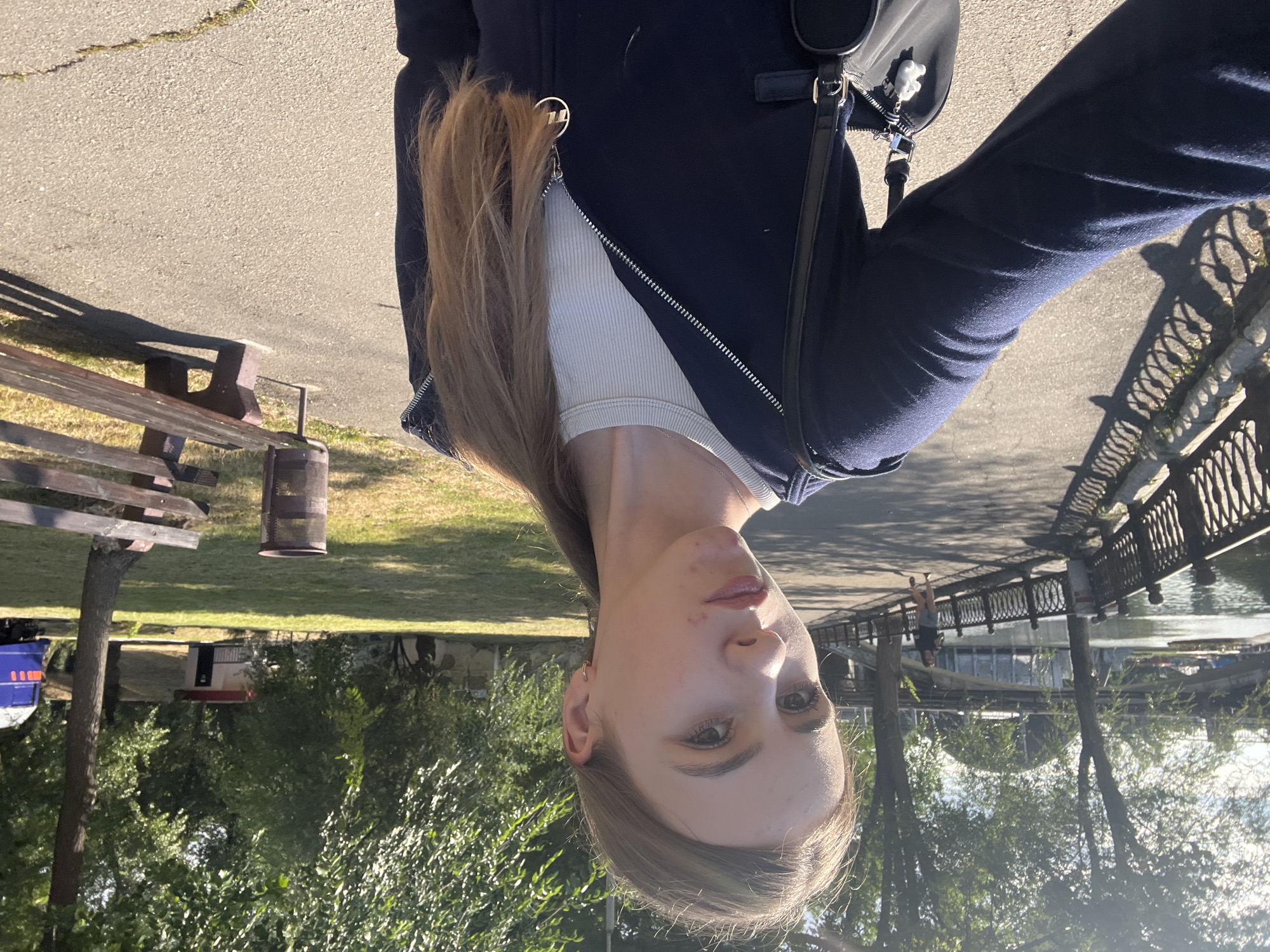 The image depicts a young woman taking a selfie outdoors in a park. She is wearing a dark blue zip-up hoodie over a white ribbed tank top and has a black purse with a white charm hanging from the strap. Her hair is long and blonde, pulled back into a ponytail.
The background suggests a park setting, possibly along a waterfront. To the left, a metal fence runs alongside a paved path. Beyond the fence, a body of water is visible, with boats and a bridge in the distance. To the right, there is a grassy area with trees, a park bench, and a trash can. A small building with a red roof is also visible in the background. The lighting indicates it is daytime, with bright sunlight casting shadows.
While the image captures a park scene, it does not contain specific landmarks that definitively place it within the city of Dnipro, Ukraine.