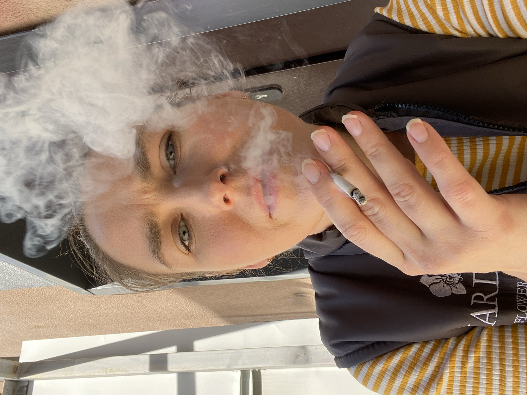 A woman is holding a cigarette and exhaling smoke. She has fair skin, light eyes, and short fingernails. She wears a black jacket over a yellow and white striped shirt. The jacket has the word "ART" and "FLOWER" embroidered on it. The background is indistinct, with a mix of brown, gray, and white surfaces. The photo was taken outdoors in daylight. There are no clear indications of the city of Yekaterinburg.