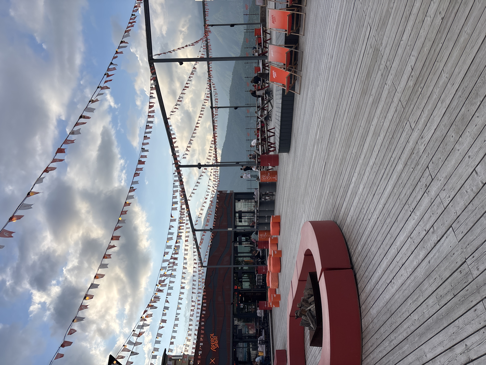 The image depicts an outdoor seating area at a venue in Estosadok, Russia, with a backdrop of rolling mountains under a cloudy sky. The foreground is dominated by a weathered wooden deck with a circular, bright red seating area containing a metal fire pit filled with logs. To the right, several red deck chairs with the "Aperol Spritz" logo are arranged. Further back, there's a building with a dark facade and a neon "Aperol Spritz" sign. The area is decorated with numerous strings of small red and white triangular flags and lights, stretching across the scene. Several groups of people are visible, some seated at tables or on benches, while others are standing. The overall impression is one of a contemporary outdoor bar or restaurant setting designed for relaxation and socializing, set against a natural mountain landscape.