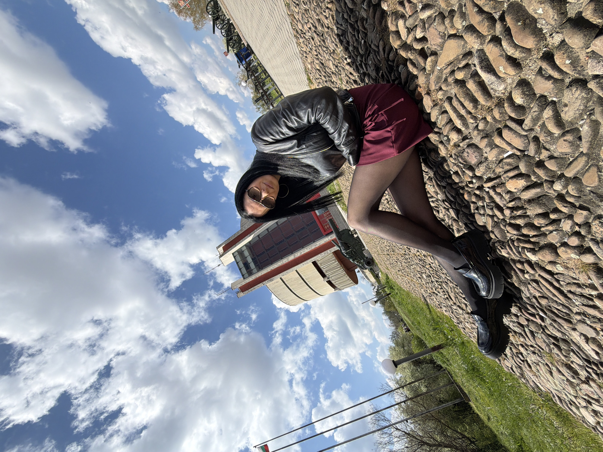 A woman in sunglasses, a black leather jacket, a maroon skirt, and black tights and shoes is posing outdoors on a sunny day. She is sitting on a cobblestone surface. Behind her is a building with a cylindrical section, suggestive of the Pleven Panorama in Pleven, Bulgaria. The sky above is blue with scattered white clouds. To the left are trees, grass, and streetlights. In the background, some bicycles are parked along a walkway. The cannons and flags confirm the building is the Pleven Panorama.