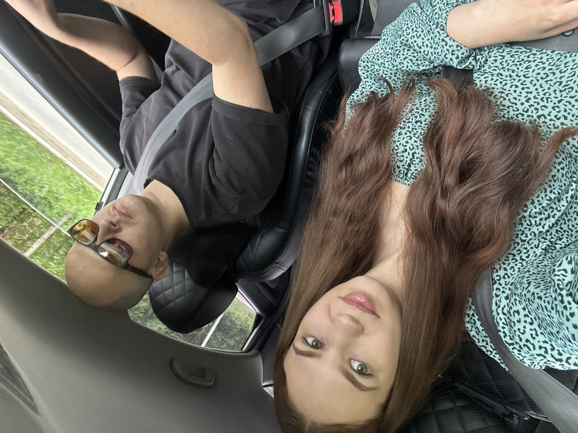 Two individuals, a man and a woman, are seated inside a vehicle. The man, with a shaved head and wearing sunglasses and a black t-shirt, is in the driver's seat. The woman, with long brown hair and wearing a teal and black leopard-print dress, is in the passenger seat. Both are wearing seatbelts. The view outside the car window shows trees and a road, suggesting they are on a journey. The image appears to have been taken from the passenger seat looking towards the driver and out the window.