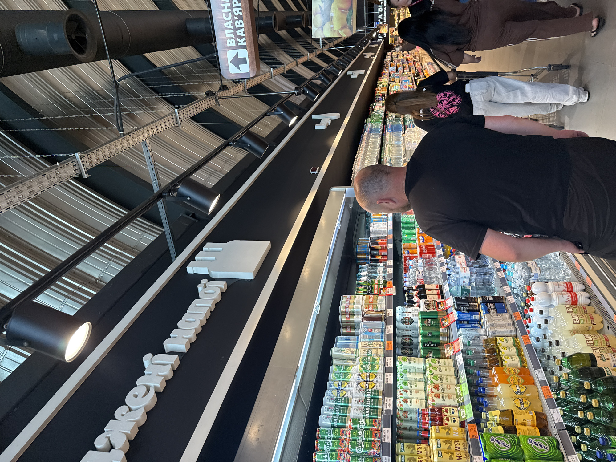 The image depicts the interior of a supermarket in Dnipro, Ukraine. In the foreground, a man with a shaved head wearing a black t-shirt walks away from the camera. Behind him, a woman with long brown hair, wearing a black t-shirt with a pink graphic and white pants, pushes a shopping cart. To the right of her, a woman in a brown tracksuit walks towards the right of the frame. The supermarket shelves are stocked with various beverages, including cans of beer and bottles of water and soda. Above the shelves, a prominent white sign with stylized text and a silhouette of buildings is mounted on a black wall. The ceiling features exposed industrial elements, including lighting fixtures, ventilation ducts, and metal conduits. A directional sign with an arrow and text in Cyrillic script is visible further down the aisle. The overall scene is brightly lit, suggesting an indoor commercial environment.