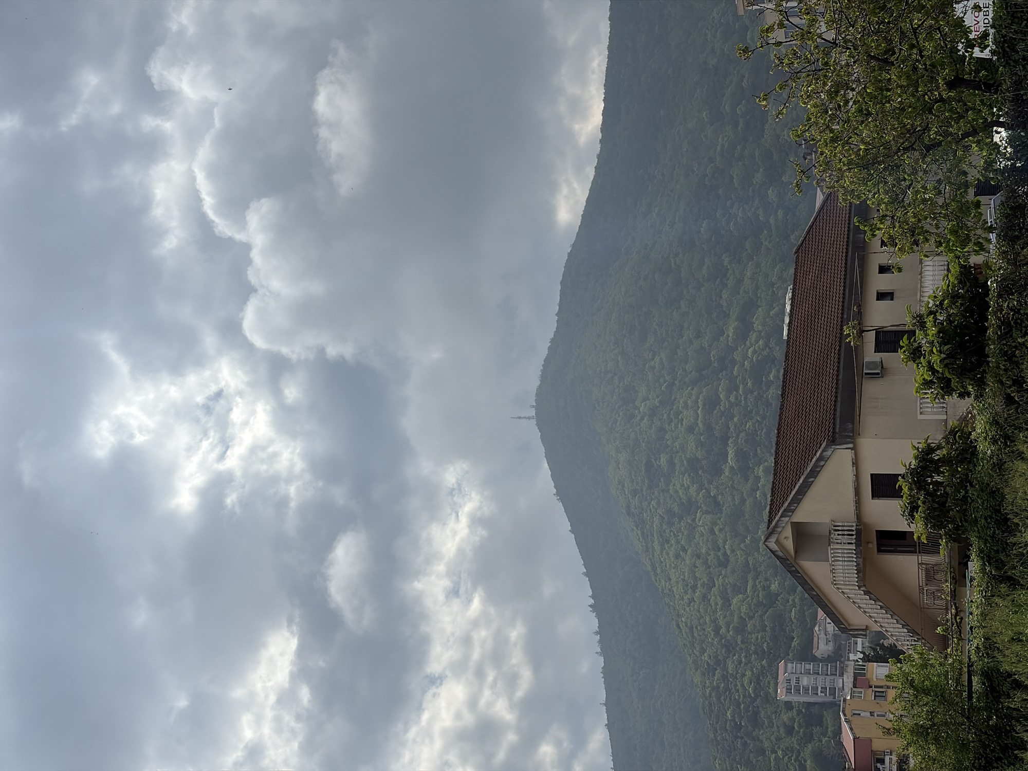 The image shows a view in Budva, Montenegro. In the foreground, a light-colored building with a reddish-brown tile roof sits on a hillside. Green foliage surrounds the building. Below, other residential buildings are visible, nestled amidst greenery. In the background, a large, densely forested mountain looms, partially obscured by mist. The sky is overcast with heavy, grey clouds dominating the scene. In the distance, a faint radio tower can be seen on top of the mountain.