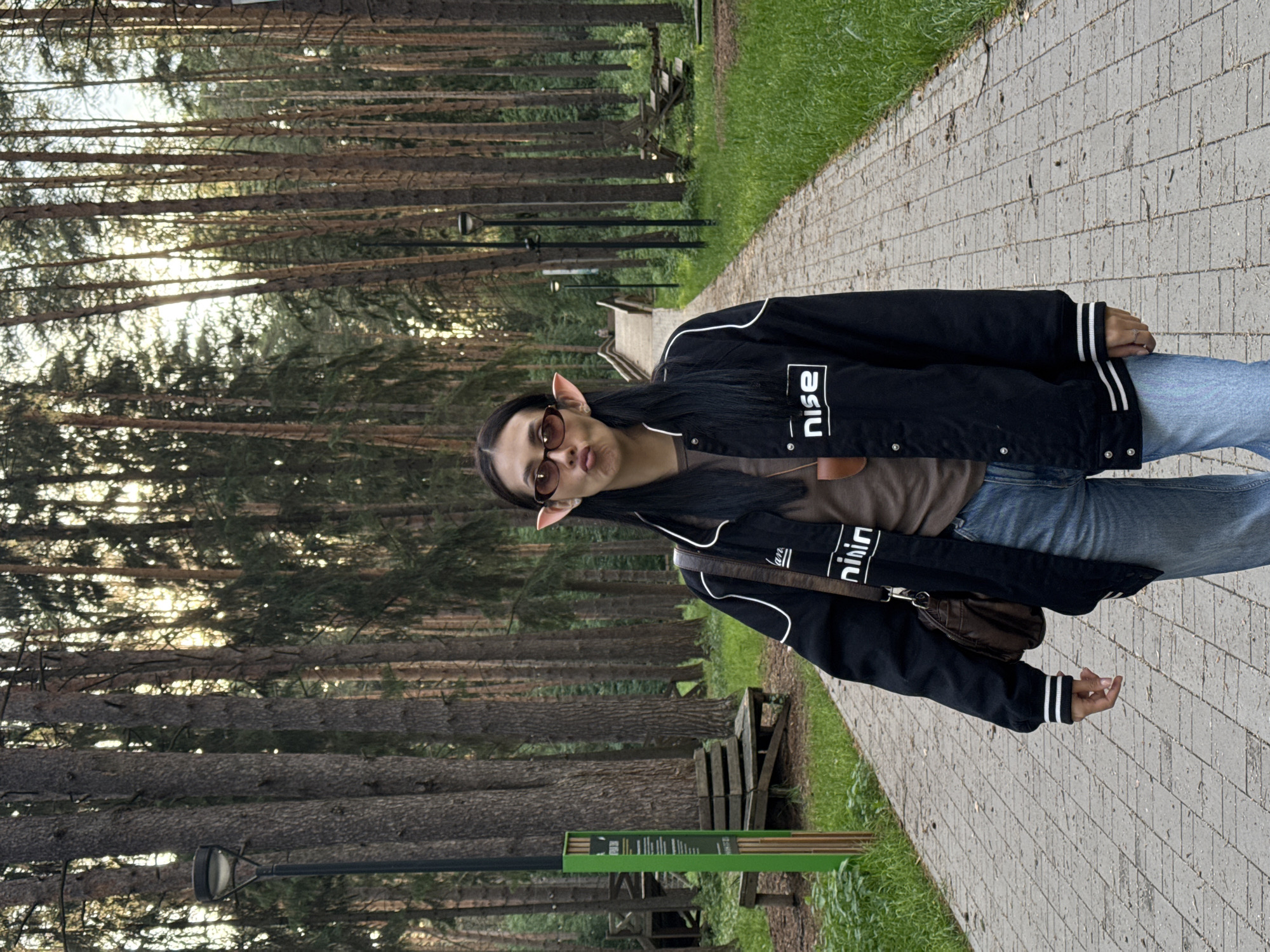 A young woman stands on a brick walkway in a wooded area. She is wearing sunglasses, a brown t-shirt, a black jacket with white lettering and stripes, blue jeans, and has a brown purse slung over her shoulder. She also has pointed ears. The wooded area consists of tall, slender trees and some grassy patches. A bench and a green sign are visible near the walkway. The sun is setting or rising, casting a soft light through the trees. The location is likely a park or recreational area within the city of Votkinsk, Russia.