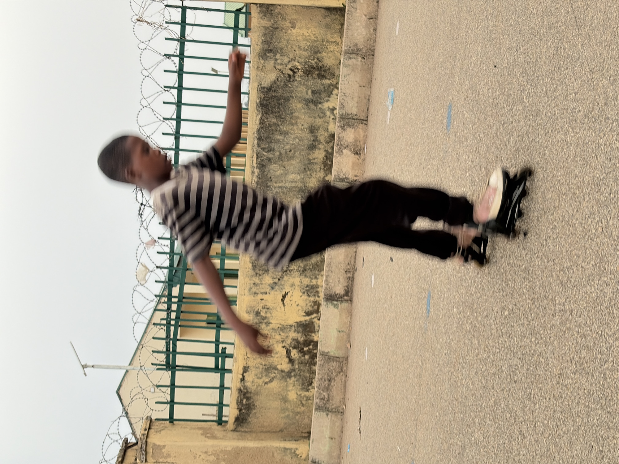 A person rollerblades on a paved area with a building featuring a green metal fence and barbed wire in the background, potentially in the city of Jos, Nigeria. The person wears a horizontally striped shirt and dark pants.