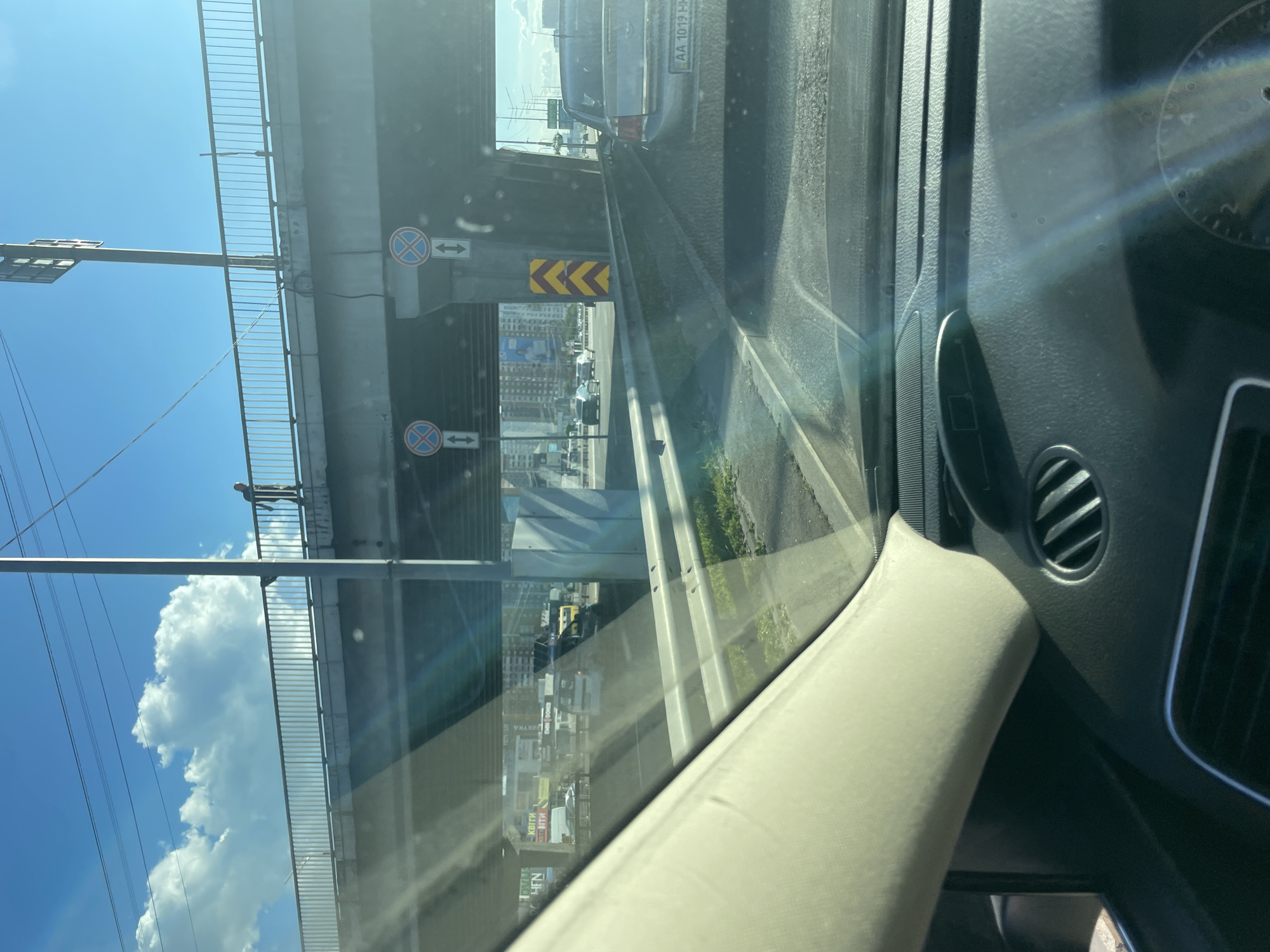 An outdoor, eye-level, medium shot shows a person standing on the railing of an overpass above a roadway in Kyiv, Ukraine. The sun is shining from the right, casting a lens flare across the image. The person is wearing dark clothing and appears to be walking along the railing. The overpass has a metal railing and is supported by concrete pillars. Below the overpass, there is a multi-lane road with several cars. In the background, there are buildings and power lines. The sky is blue with some white clouds. The scene is captured from inside a vehicle, with part of the dashboard and windshield visible in the foreground.