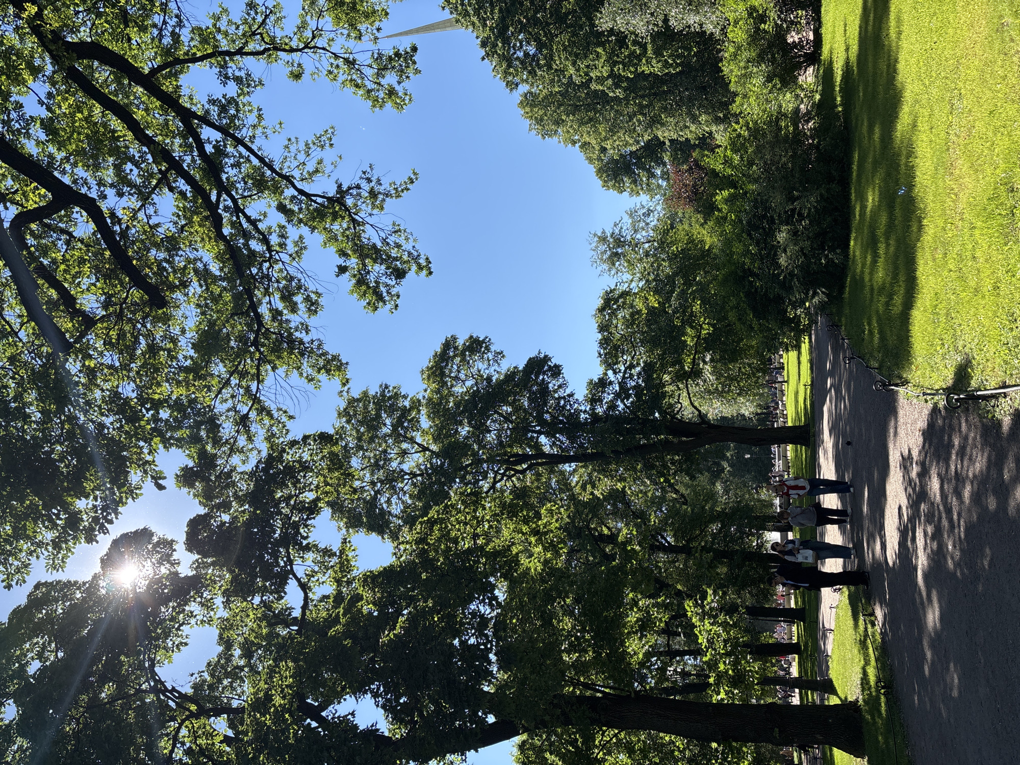 The media file depicts a sunny day in Saint Petersburg, Russia, likely during the late morning or early afternoon, given the bright sunlight and distinct shadows. The scene is an outdoor park with a paved pathway lined by lush green grass and mature trees.
Several people are visible walking along the pathway. In the foreground, three individuals are walking together. The person closest to the viewer is wearing a dark suit, and the two people behind them are dressed more casually, one in a white shirt and jeans, and the other in a grey jacket and blue jeans. Further down the path, more people can be seen in the distance, suggesting the park is a popular public space.
The trees, with their dense green foliage, cast dappled shadows across the pathway and grass, creating a visually interesting contrast between light and shade. The sky is a clear, bright blue, indicating fair weather. The sunlight is strong, with visible sun flares and rays filtering through the leaves of a large tree in the upper left portion of the image. In the background, a tall, light-colored, pointed structure can be seen emerging above the tree line, a possible landmark within the city.
There is no visible text in the media file. The overall atmosphere is peaceful and recreational, conveying a sense of enjoying a pleasant day outdoors in a well-maintained park.