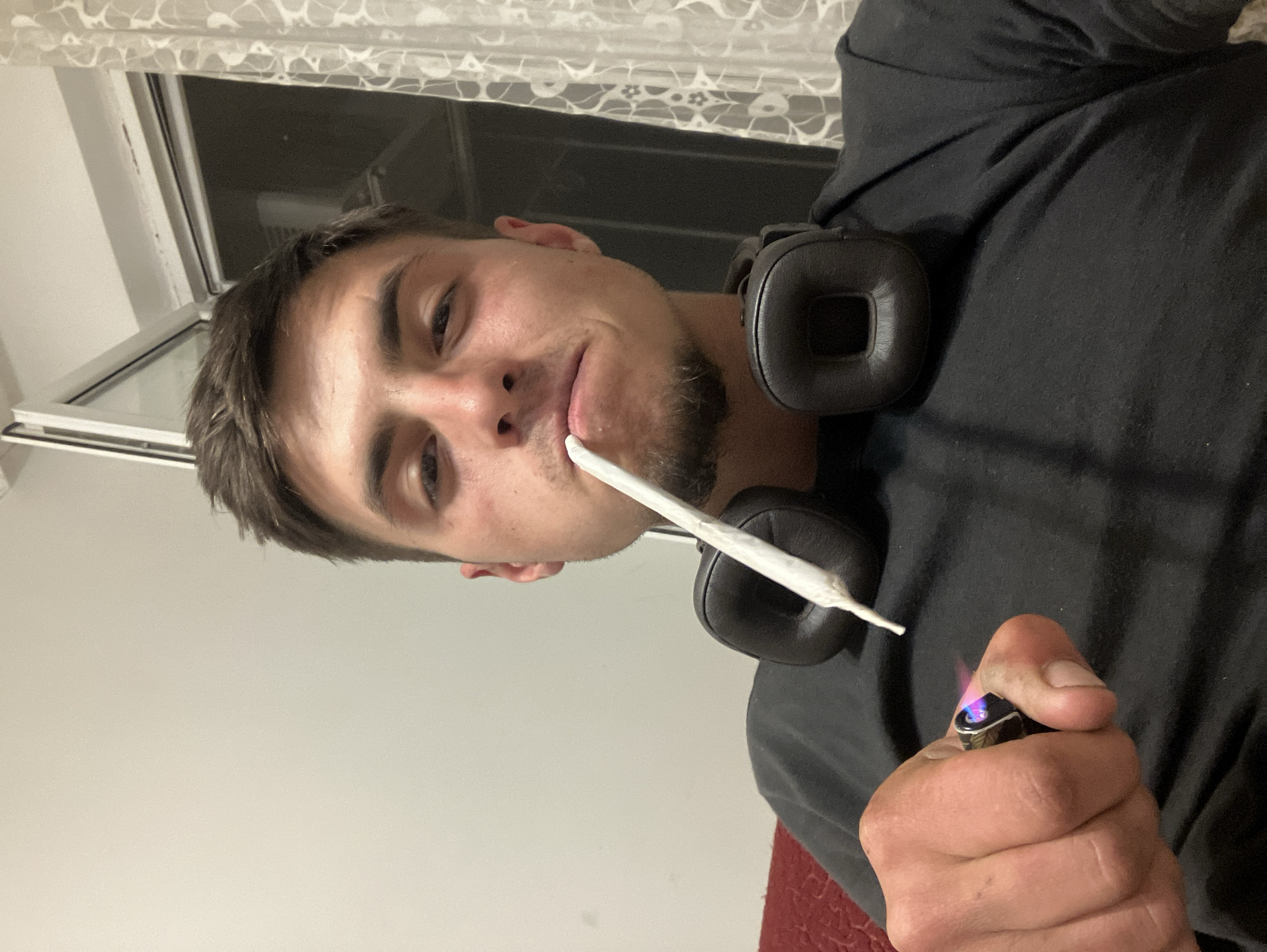 A man is holding a lighter in his right hand with the flame lit. A rolled cigarette is in his mouth, and he is wearing black headphones around his neck. The background includes a window with a white curtain and a gray wall. The location appears to be indoors. I cannot determine the city of Plovdiv, Bulgaria as it is not apparent from the image.