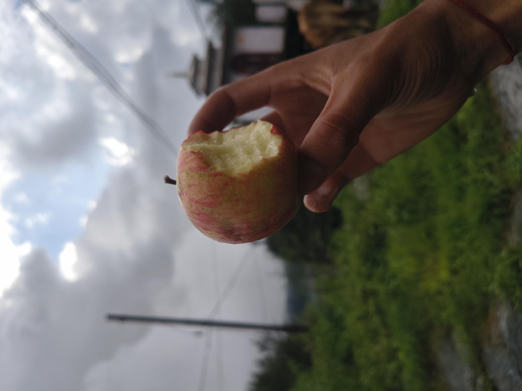 A person's hand holds a partially eaten apple in front of a cloudy sky. The apple is red and yellow with visible bite marks revealing the white flesh. The background is blurred, showing green foliage, a blurry building structure, and what appears to be a cow. Power lines are visible against the sky. The scene appears to be outdoors in a rural or semi-rural setting.