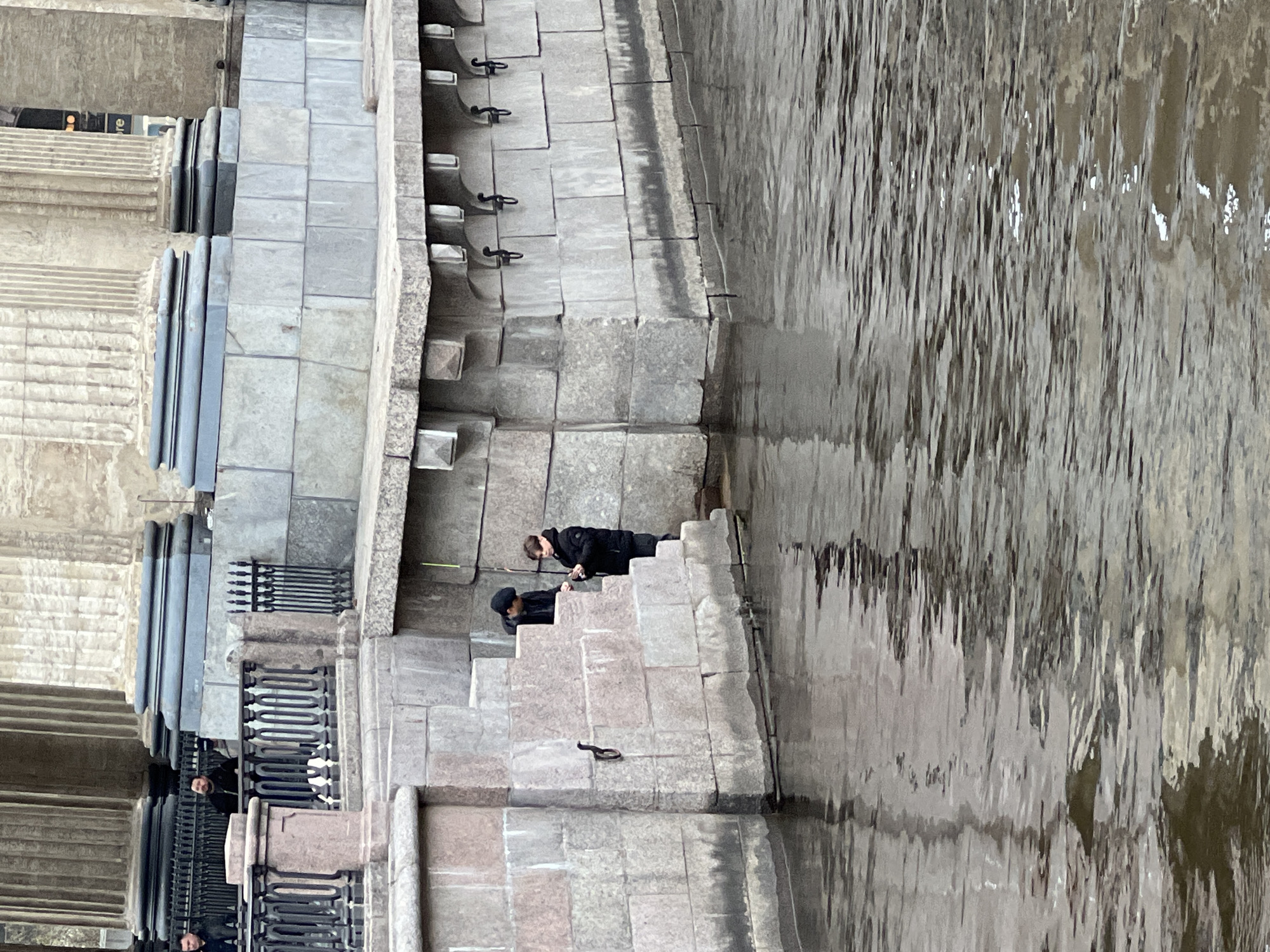 The image displays a stone embankment alongside a body of water, likely a river or canal, under overcast conditions. The perspective looks down onto a series of broad, grey stone steps that descend towards the water.
Two individuals are present on these steps, appearing to be fishing. One person, dressed in a dark jacket and trousers, stands on a higher step, holding a long fishing pole extended towards the water. Below them, on a lower step, a second person wearing a dark hat and dark clothing is kneeling or crouching, also with a fishing pole directed into the water.
The embankment is constructed from large, robust stone blocks. Along the edge of the water, black metal mooring rings or hooks are integrated into the stonework. The water itself is a murky greyish-brown, reflecting the muted light of the sky and showing ripples across its surface.
In the background, behind the fishing activity, more elaborate stone architecture is visible, including columns and intricate black metal railings, suggesting grand city buildings. The overall scene conveys a calm, possibly cool, atmosphere. No significant text is clearly visible, though a faint, partial "re" can be barely discerned on a distant dark surface in the upper left of the original image.